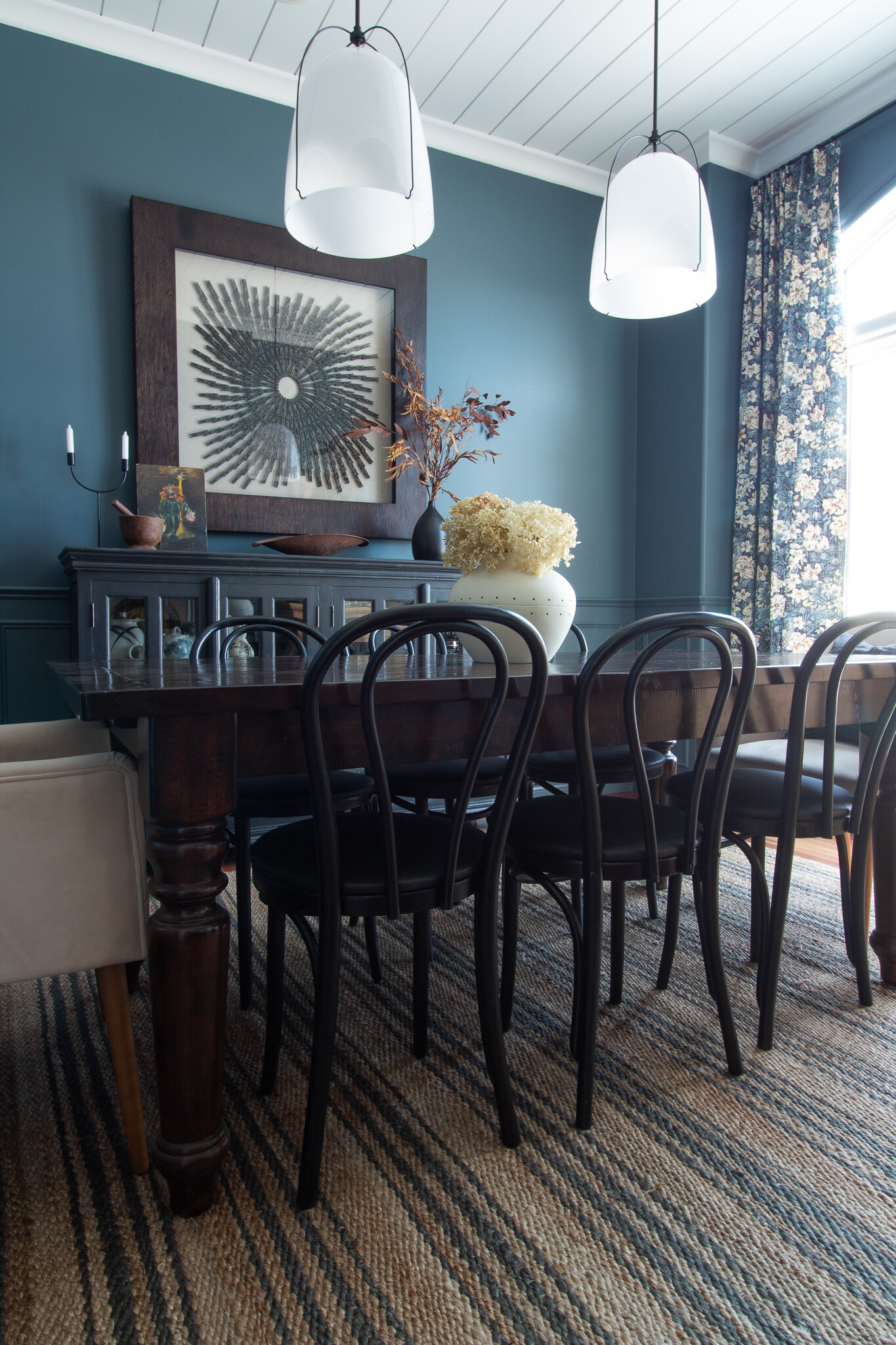 Blue cdinging room with white rejuvination pendants and a white shiplap ceiling, box trim wainscotting, floral curtians, black bentwood chairs, dark wood table, black sideboard, clothespin art, jute striped rug