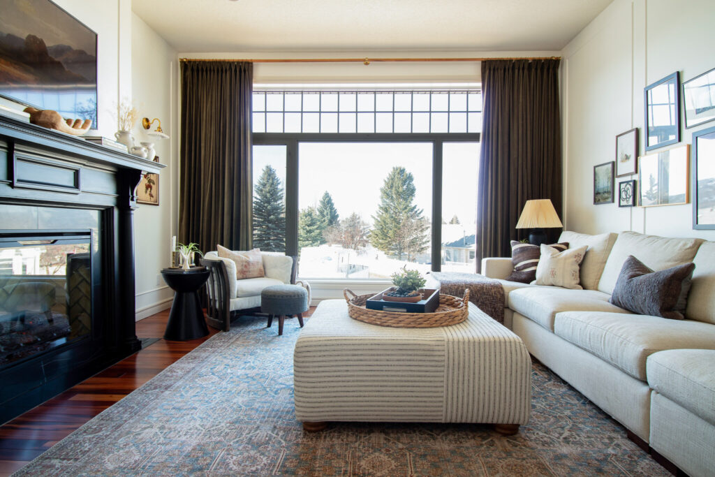 Living room with large window with green velvet drapes, beige sectional, upholstered striped ottoman, vintage style rug, black fireplace with antique mantel, vintage chair