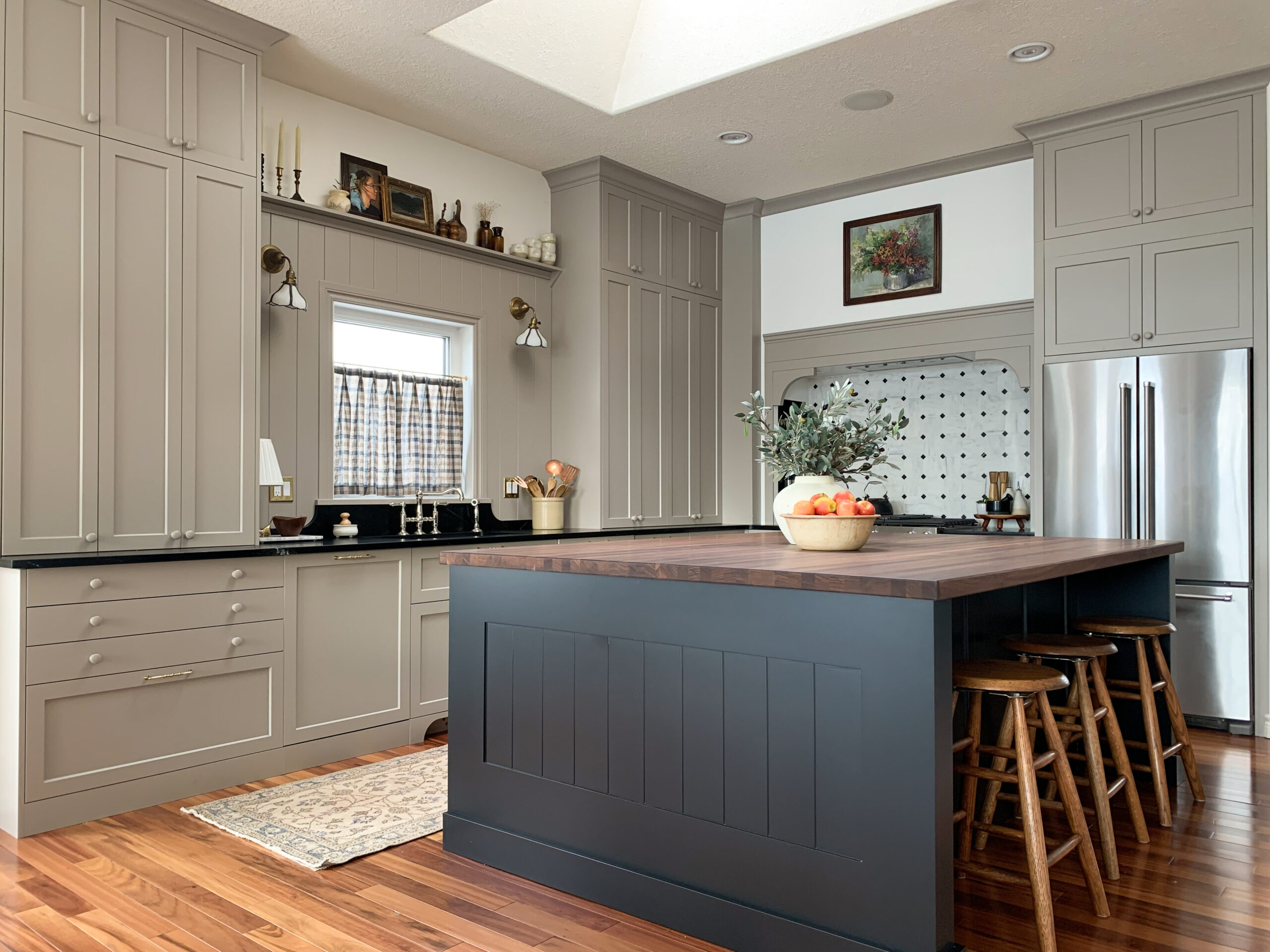 kitchen with beige cabinets, black island, black soapstone and walnut butcherblock countertops, built in stove nook with custom vent hood, marble counters, cafe curtains, and a shelf over a window