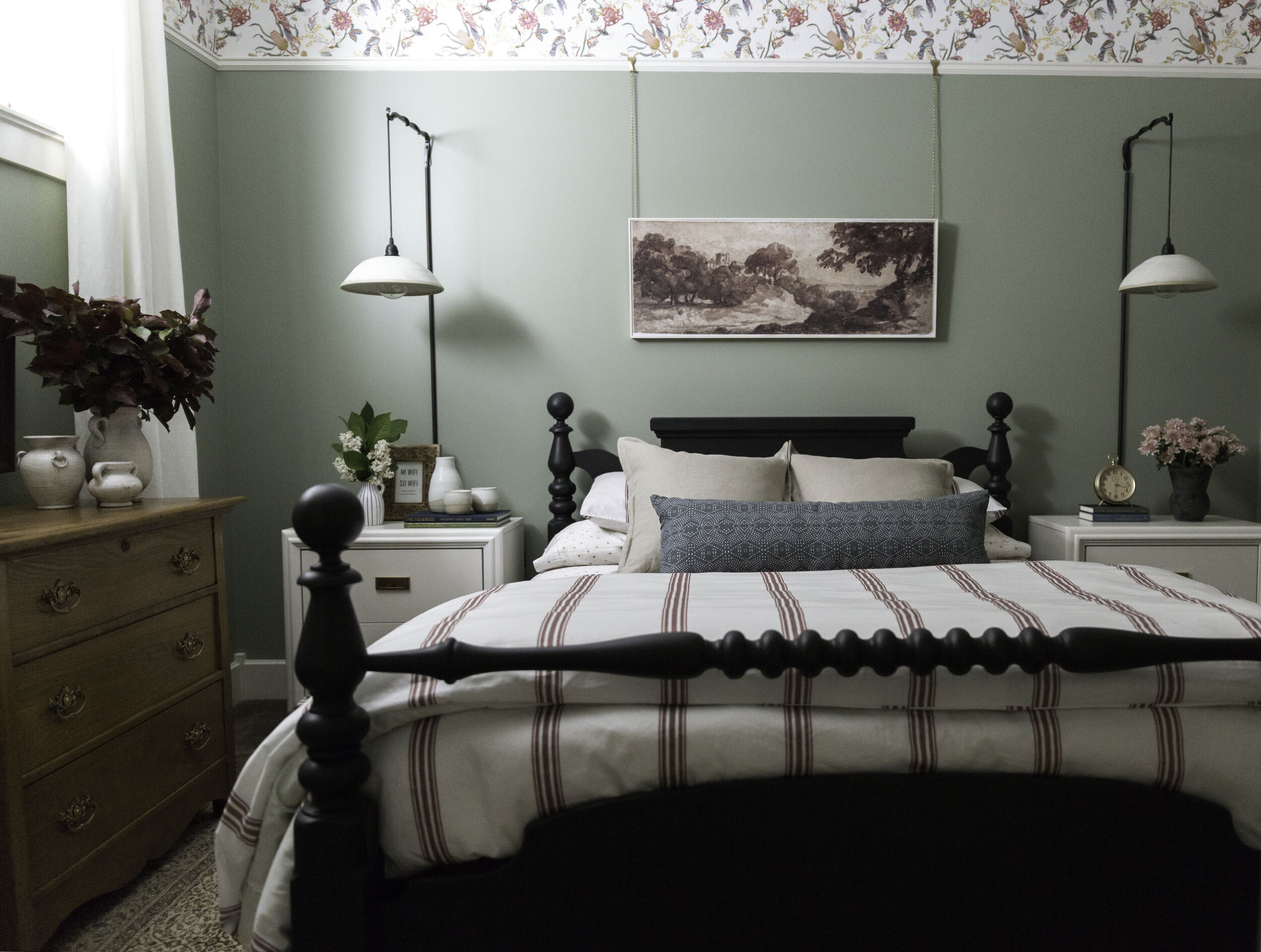green bedroom with wallpaper at top of wall, red striped bedding, white modern nightstands, hanging ceramic wall sconces, vintage rug and black antique bed and antique wood dresser