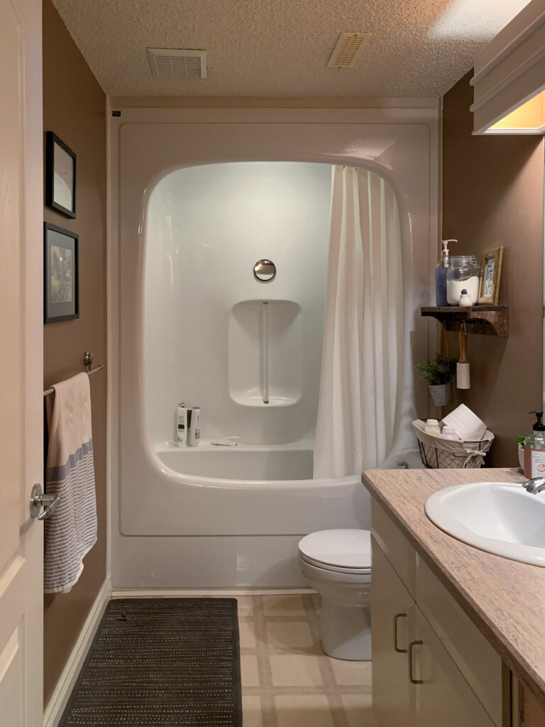 Before 90's bathroom with rounted built in tub, linoleum floors, popcorn ceiling, brown walls, white flat front cabinets and laminate counters