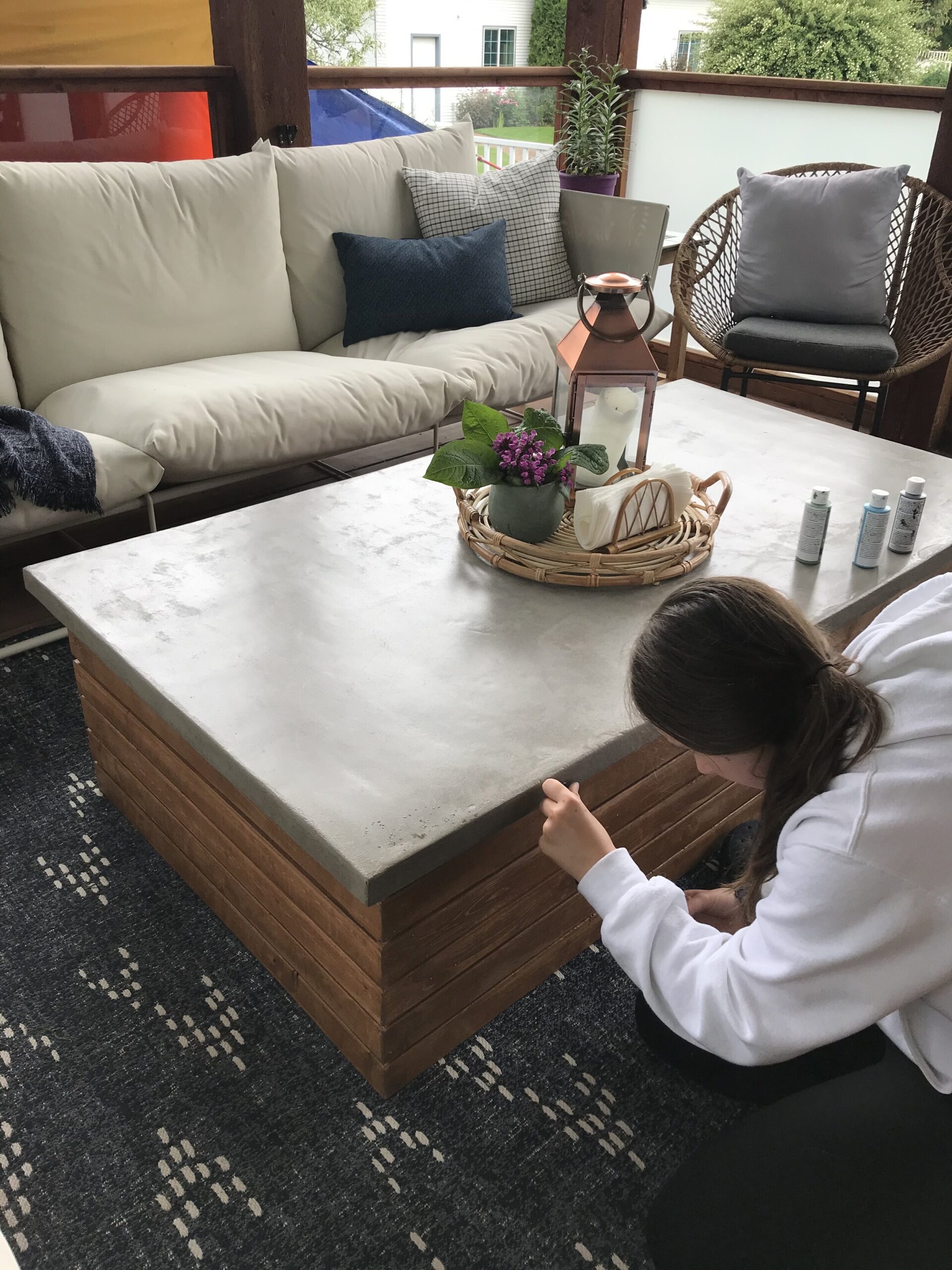 girl touching up the edges of the concrete with paint on a concrete table top on a deck with flowers and outdoor couch and blue outdoor rug