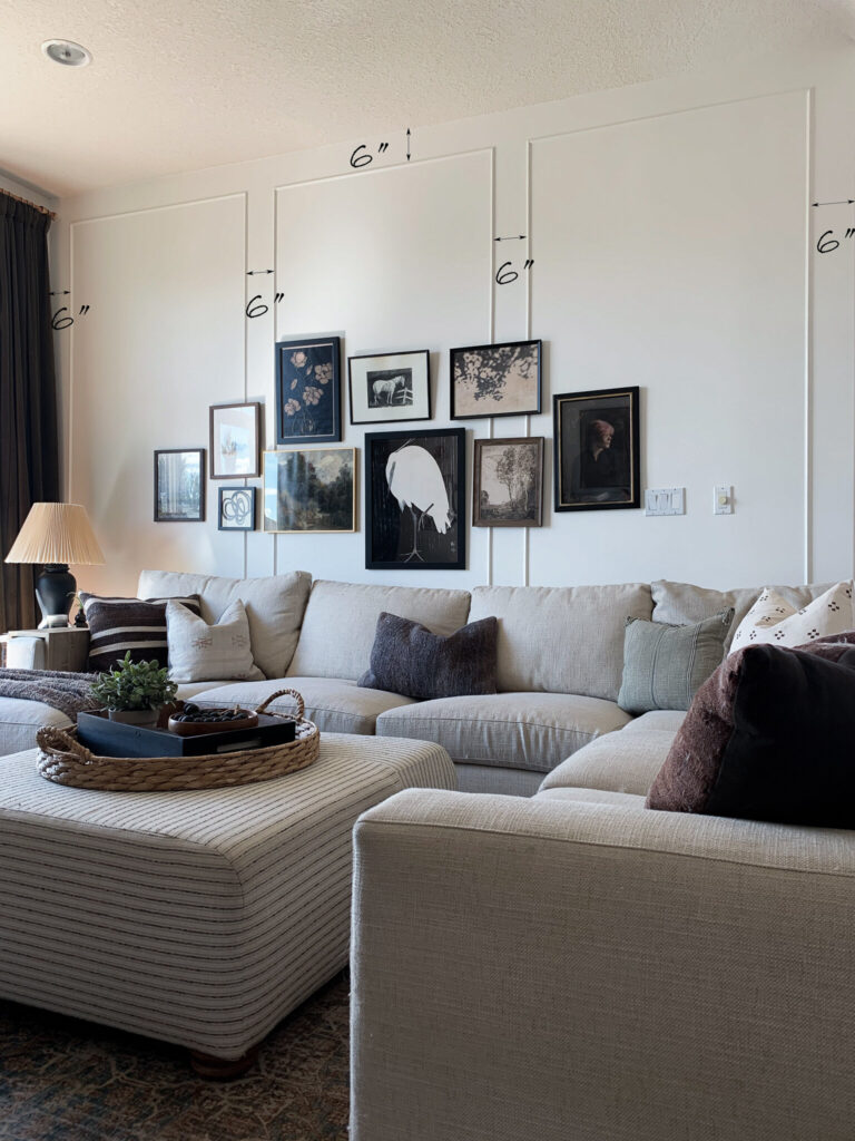 Living room showing box trim moudling installed with a gallery wall of vintage art over it, with dimensions showing distance between boxes. Living room also has a sectional and an upholstered ottoman