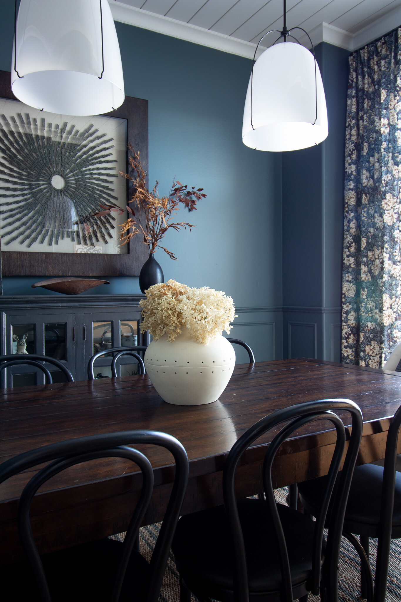 Dining room with blue walls, white shiplap ceiling, modern white rejuvenation plendants, floral curtains, jute striped area rug, black bentwood chairs, upholstered end chairs, dark wood table, black sideboard, and oversized art