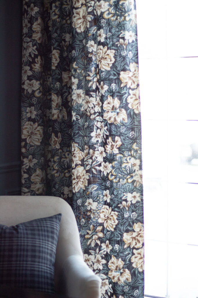 floral blue curtains with beige flowers on a widnow with white upholstered chair in front of window with blue pillow on it