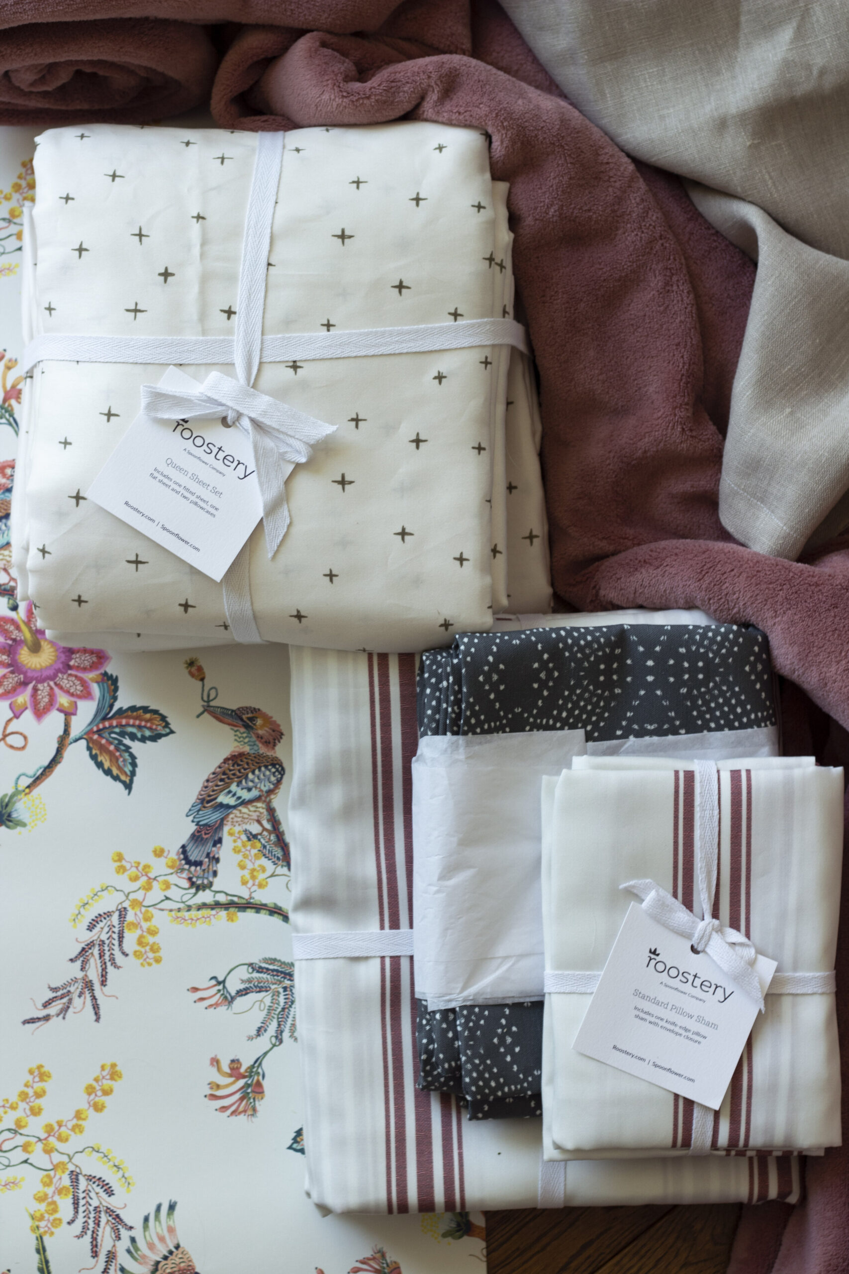 flatlay with pink bedding and beige linen, white sheets with green crosses, blue pillow, white duvet with red stripes, floral wallpaper