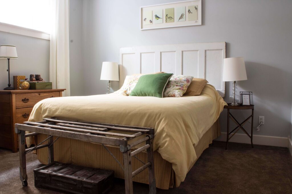 bedroom with grey walls and white trim, yellow bedding, old door as a headboard, wood dresser, white curtains and brown carpet