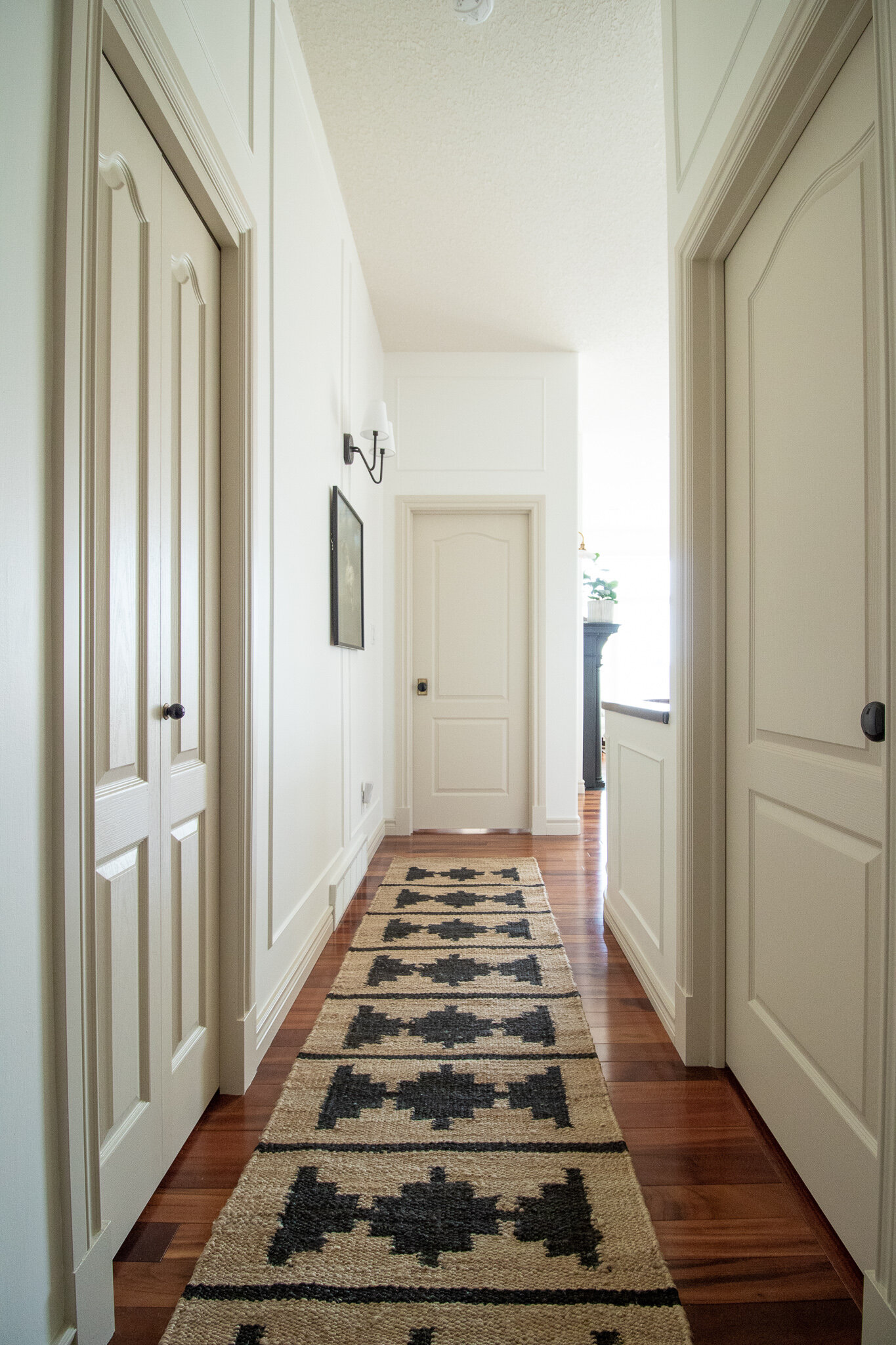 hallway with white walls with box trim moulding, beige trim and doors, black sconce, jute runner with black geometric design and brass and black doorknobs