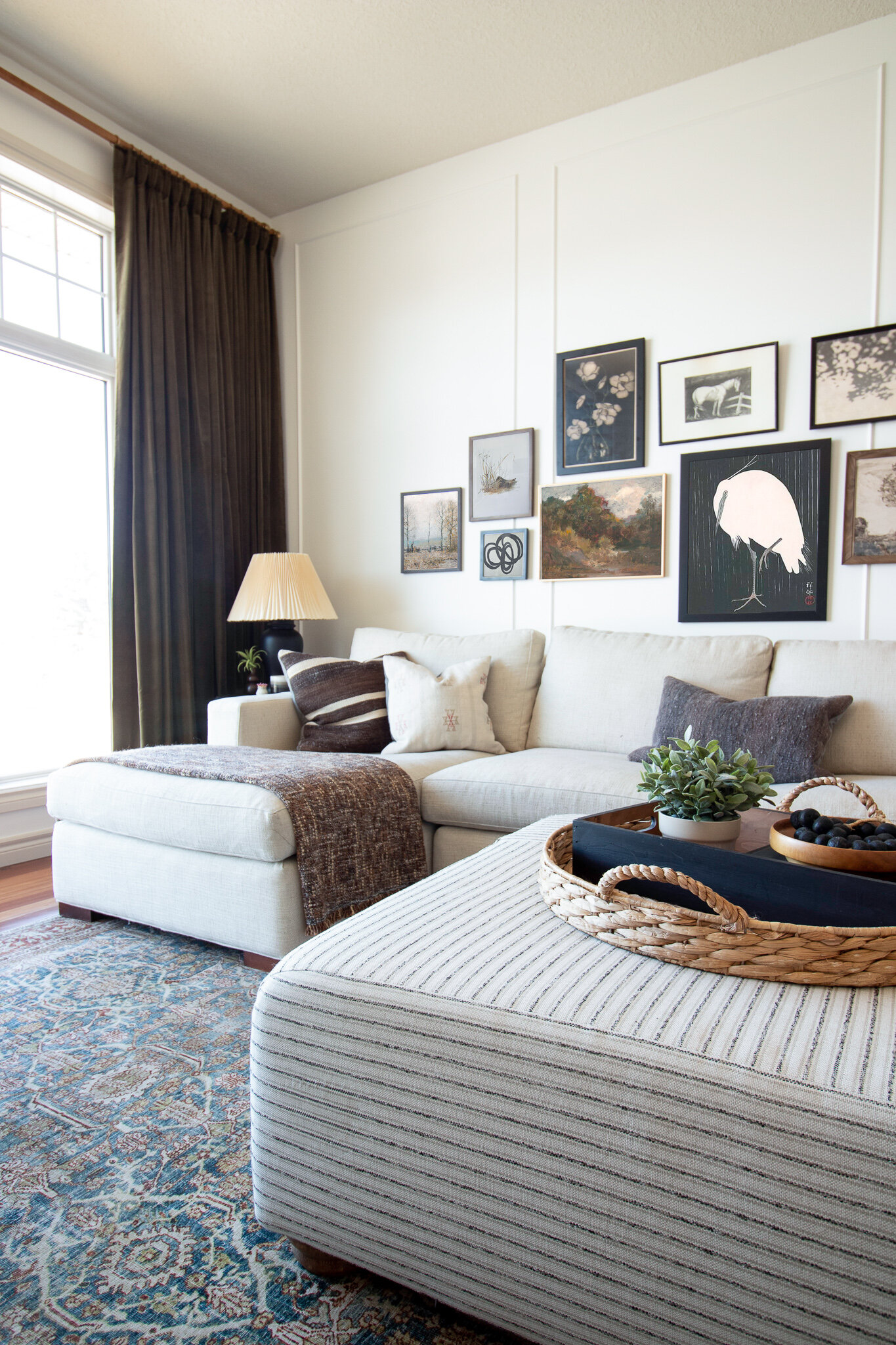 Living room with beige sectional, gallery wall of vintage art, green velvet curtains, striped upholstered ottoman, wicker tray on ottoman 