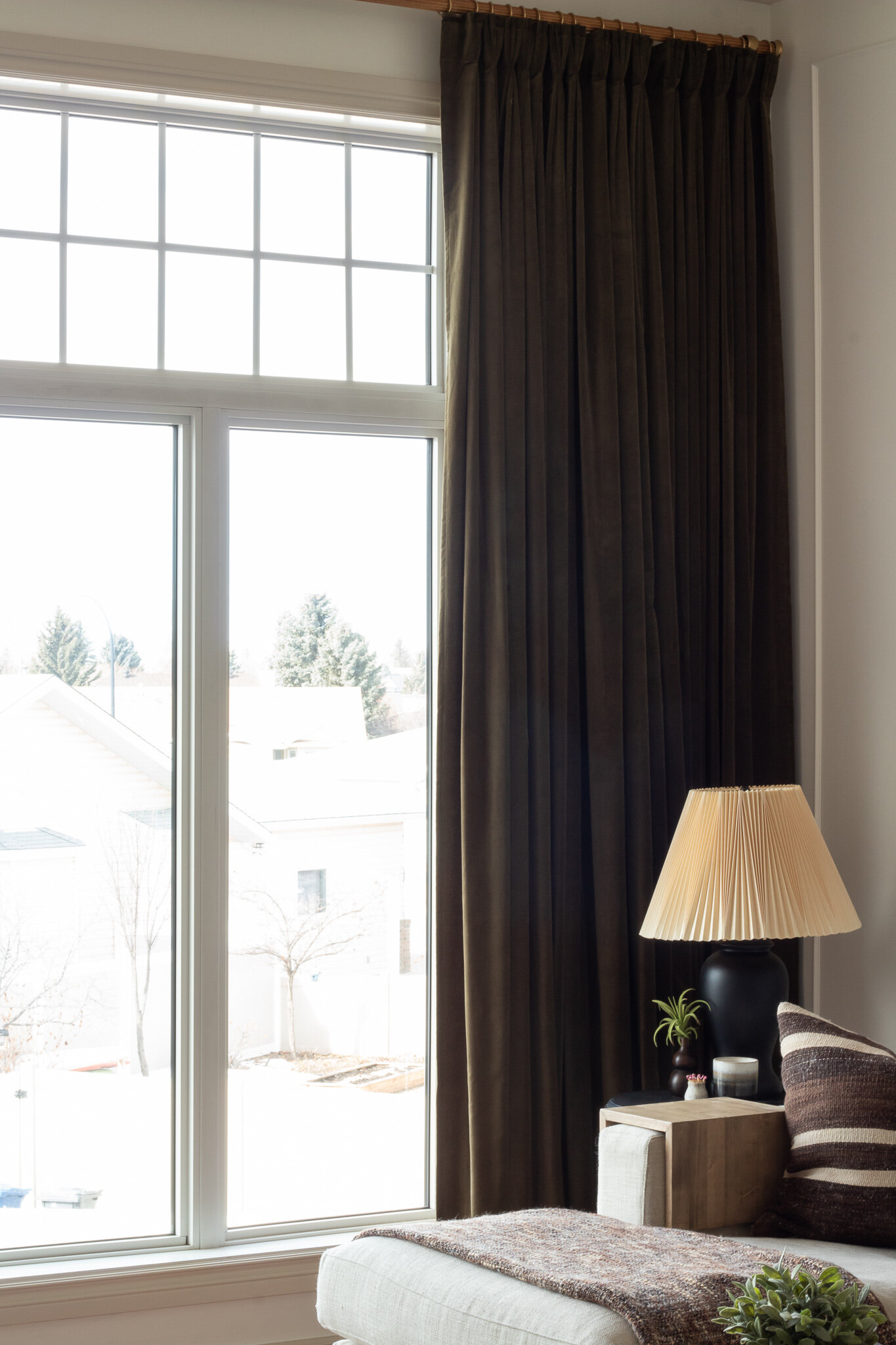 Corner of a room with large windown with green velvet curtains, corner of sectional with black lamp and pleated shade on a side table