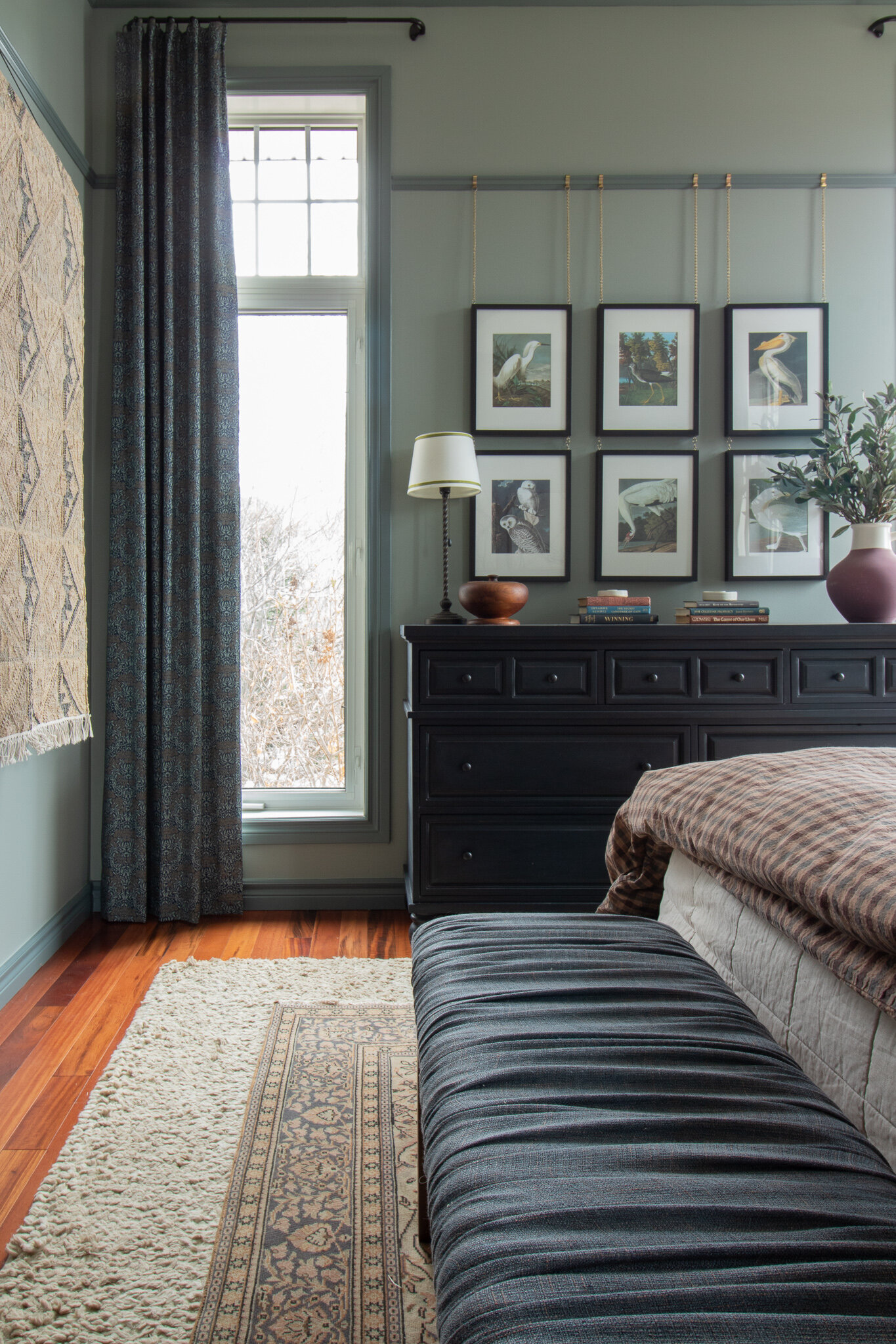 facing window with black dresser next to window . floral curtains, gallery wall of bird art hanging from picture rail, blue bench in foreground