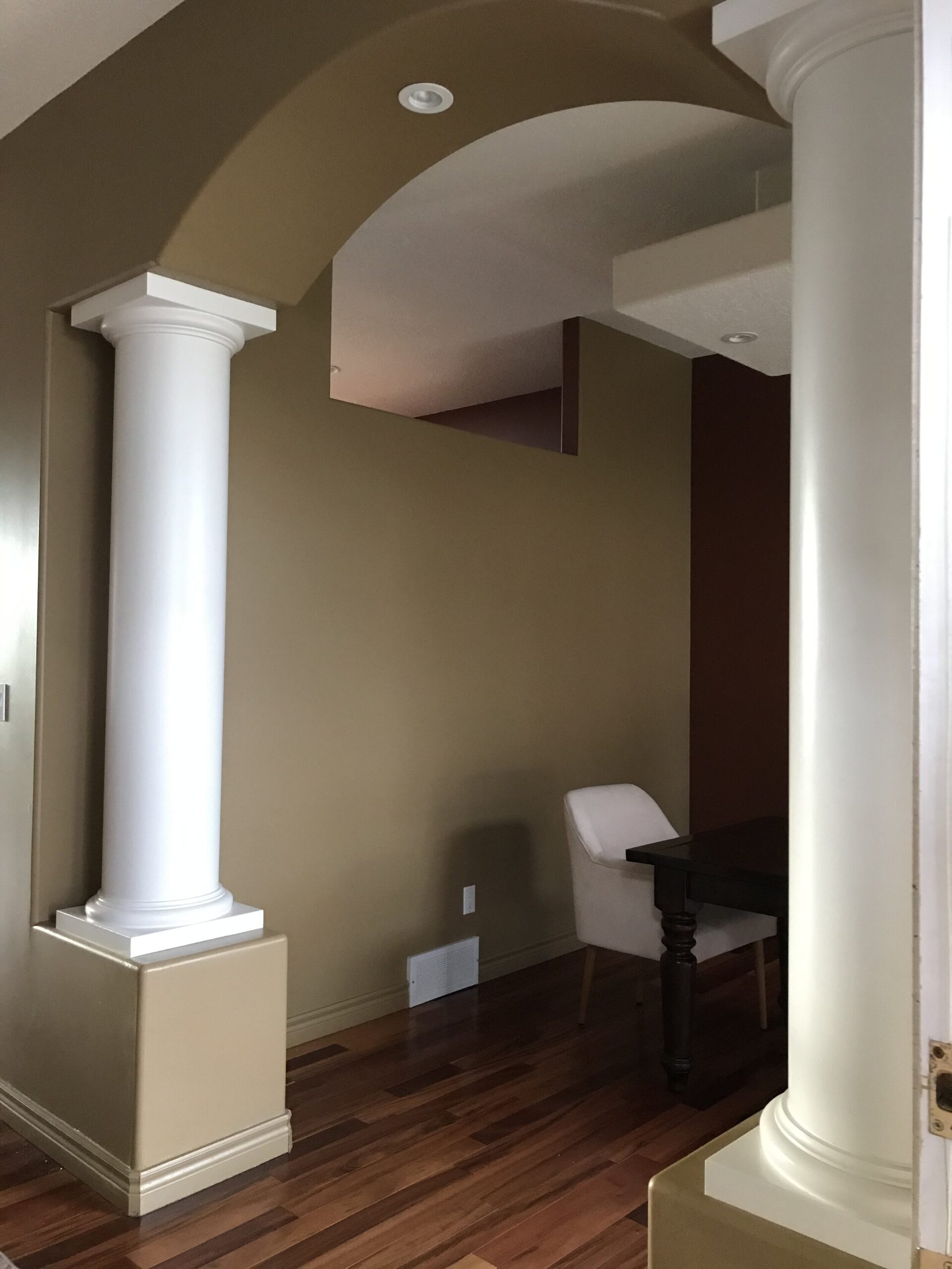 Dining room with red and brown walls, minimal furniture, large boxed light and columns framing the arched opening and a cut out in the wall at the ceiling