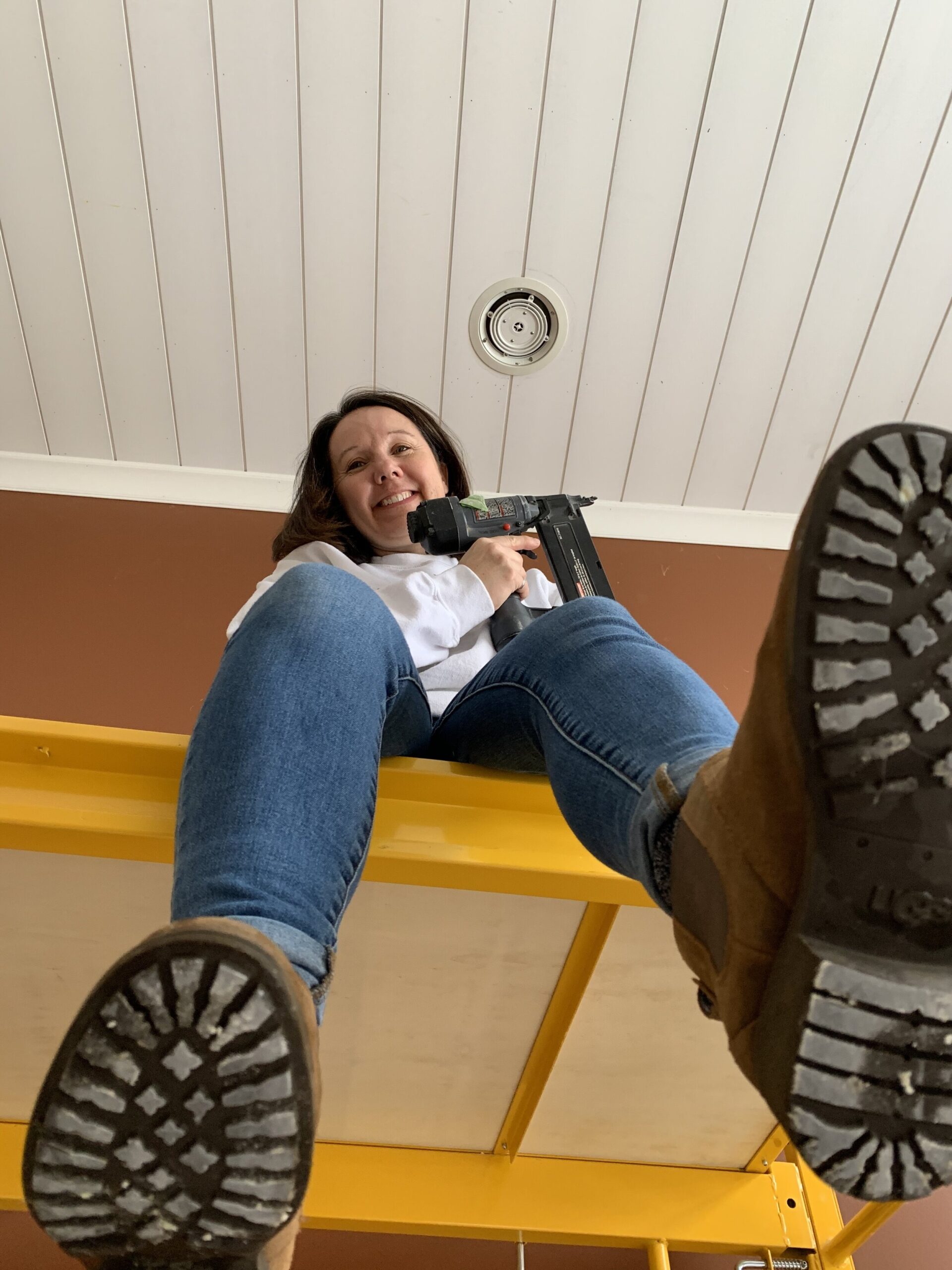 girl sitting on a scaffold holding a nail gun with a shiplap planked white ceiling in background