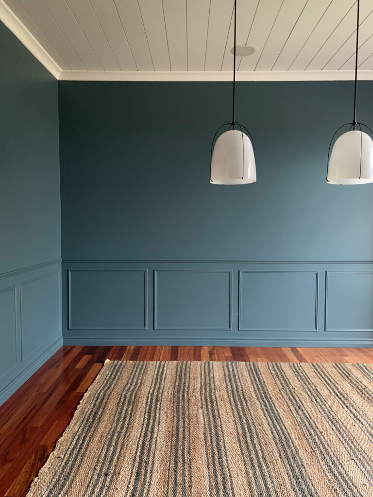 Dining room with no furniture but a jute striped rug, blue walls with wainscotting, white shiplap ceiling and white rejuvination pendants