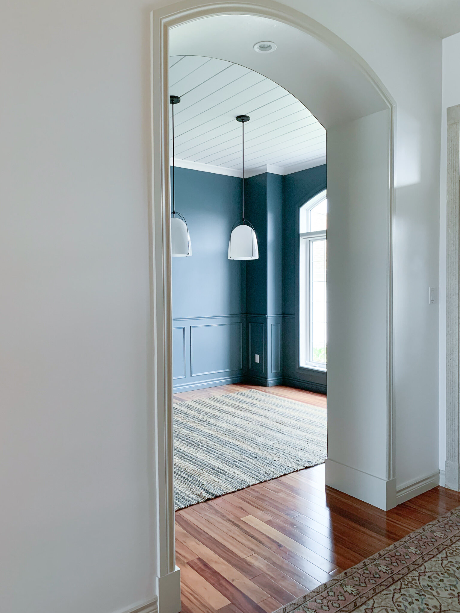 Arched opening into dining room with blue walls, box trim paneling, shiplap ceiling, white glass pendant lights