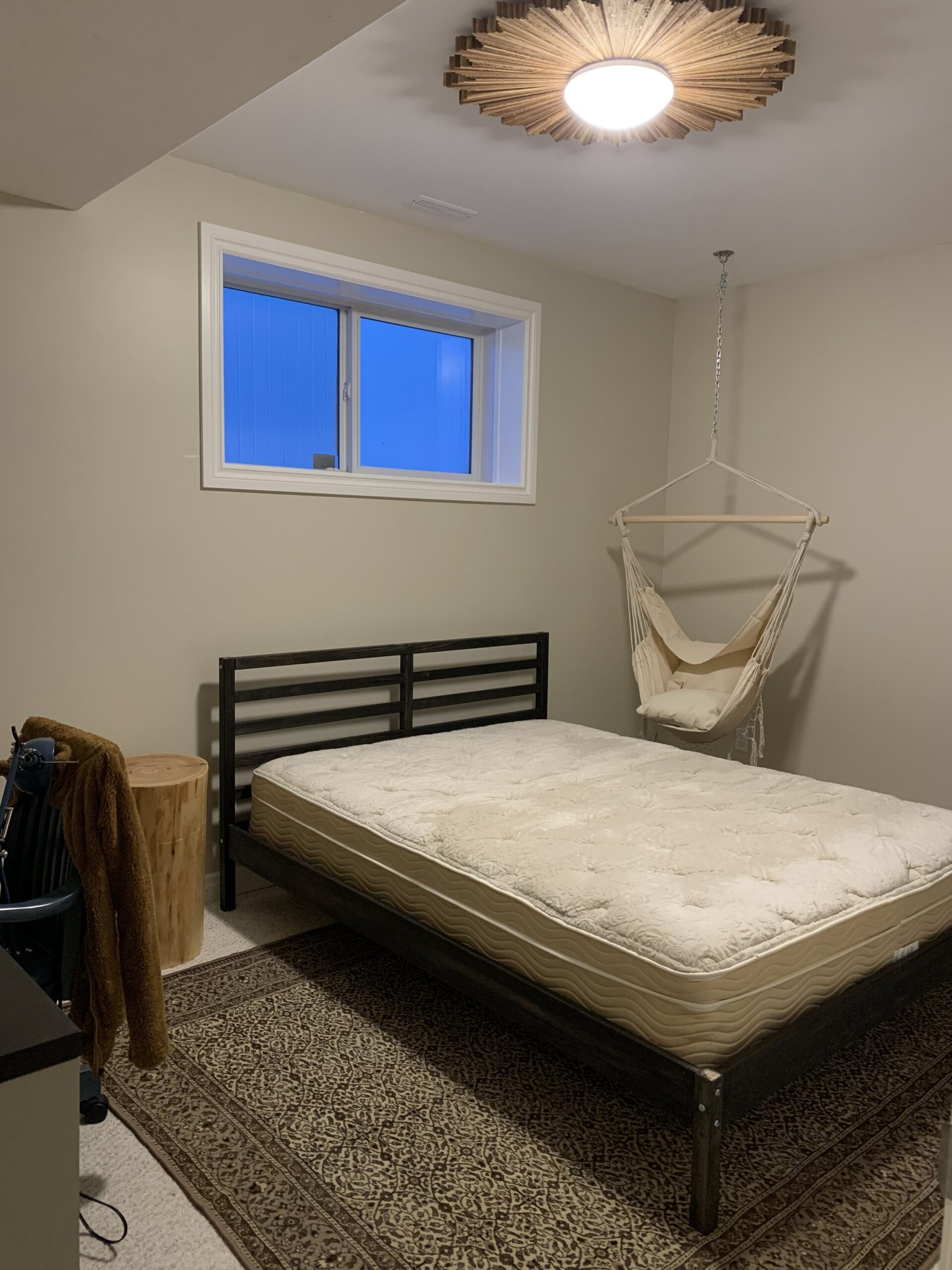 bedroom with beige walls and white trim, hanging macrame chair, black wood bed and plain mattress, starburst wood ceiling light and brown vintage rug