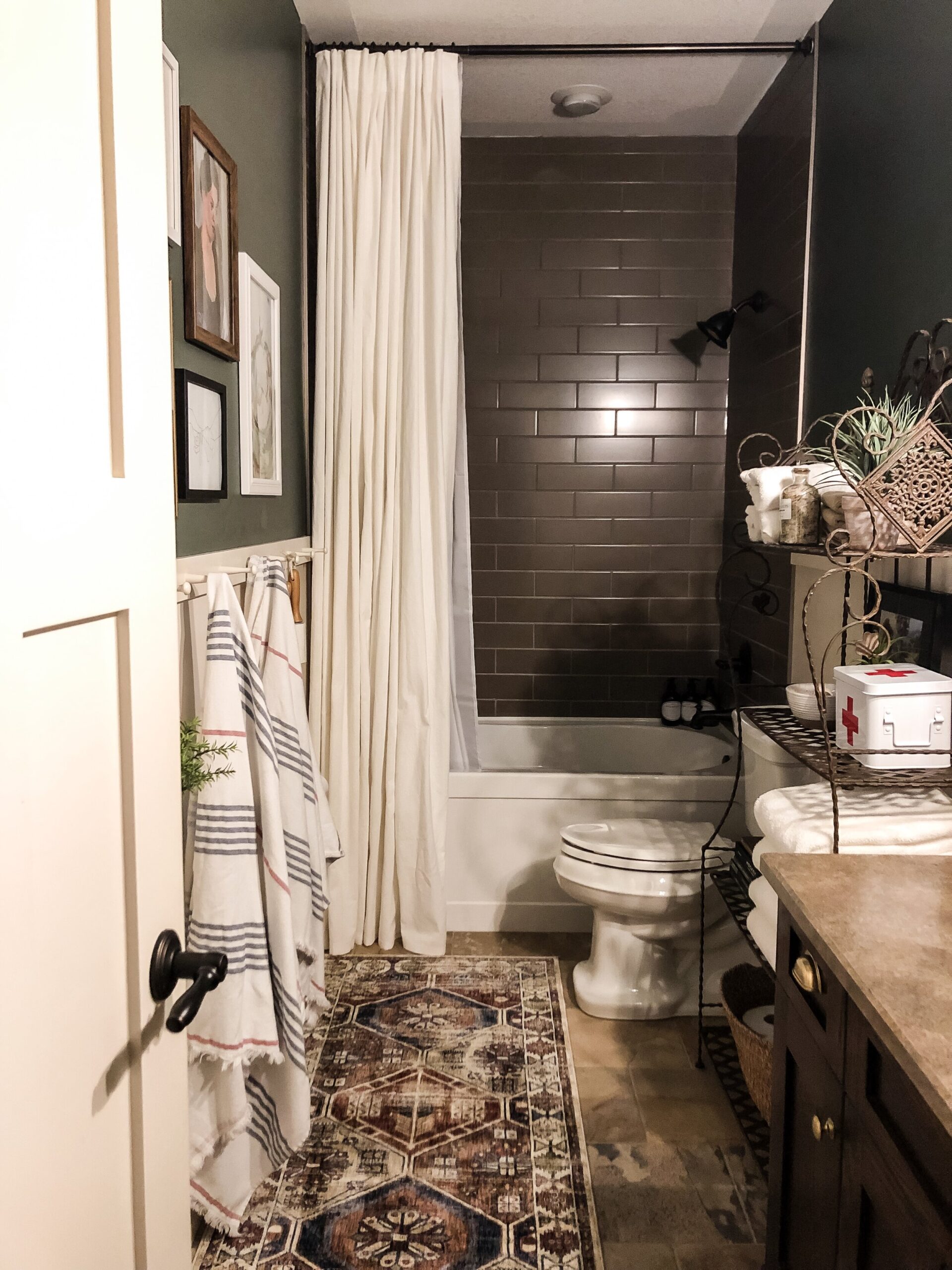 Full length shower curtain in white with brown subway tile and green walls with white wainscotting and a vintage style rug
