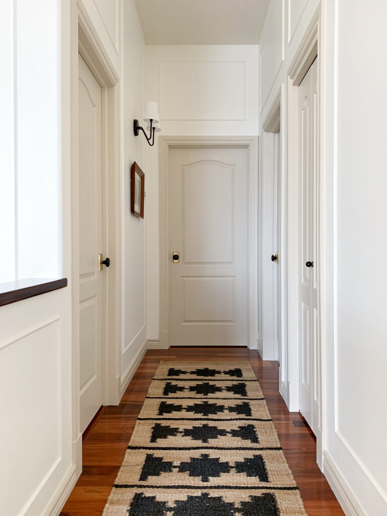 a hallway with many doors with white walls and beige trim and doors with a jute runner showing box trim moulding installed above a door offset from the door casing