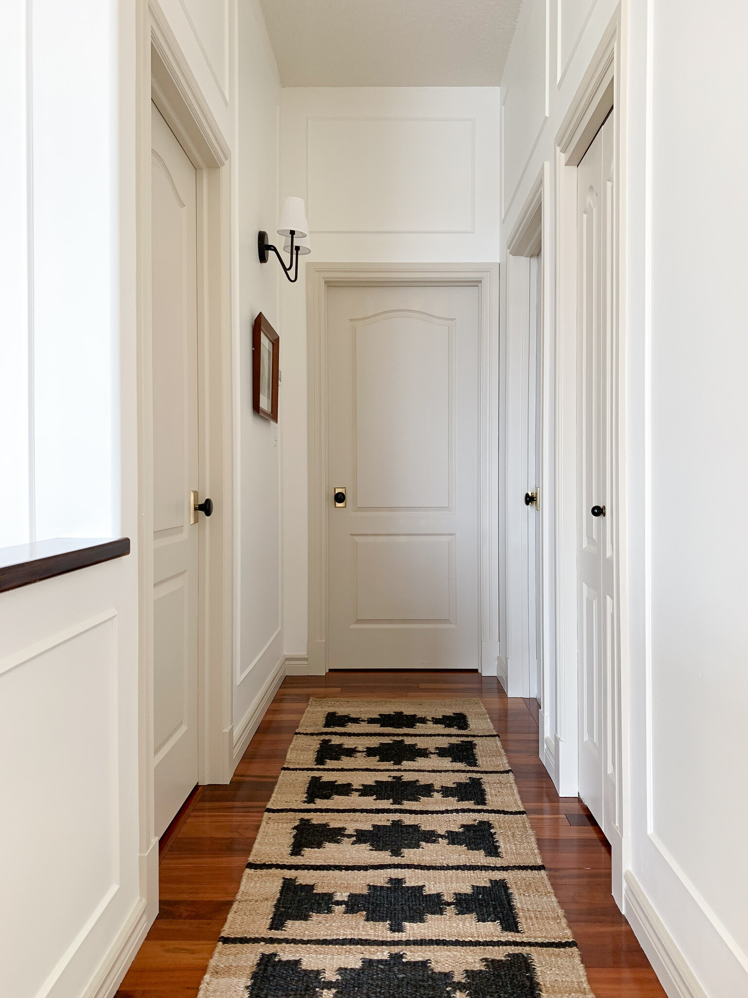 a hallway with many doors with white walls and beige trim and doors with a jute runner showing box trim installed above a door offset from the door casing