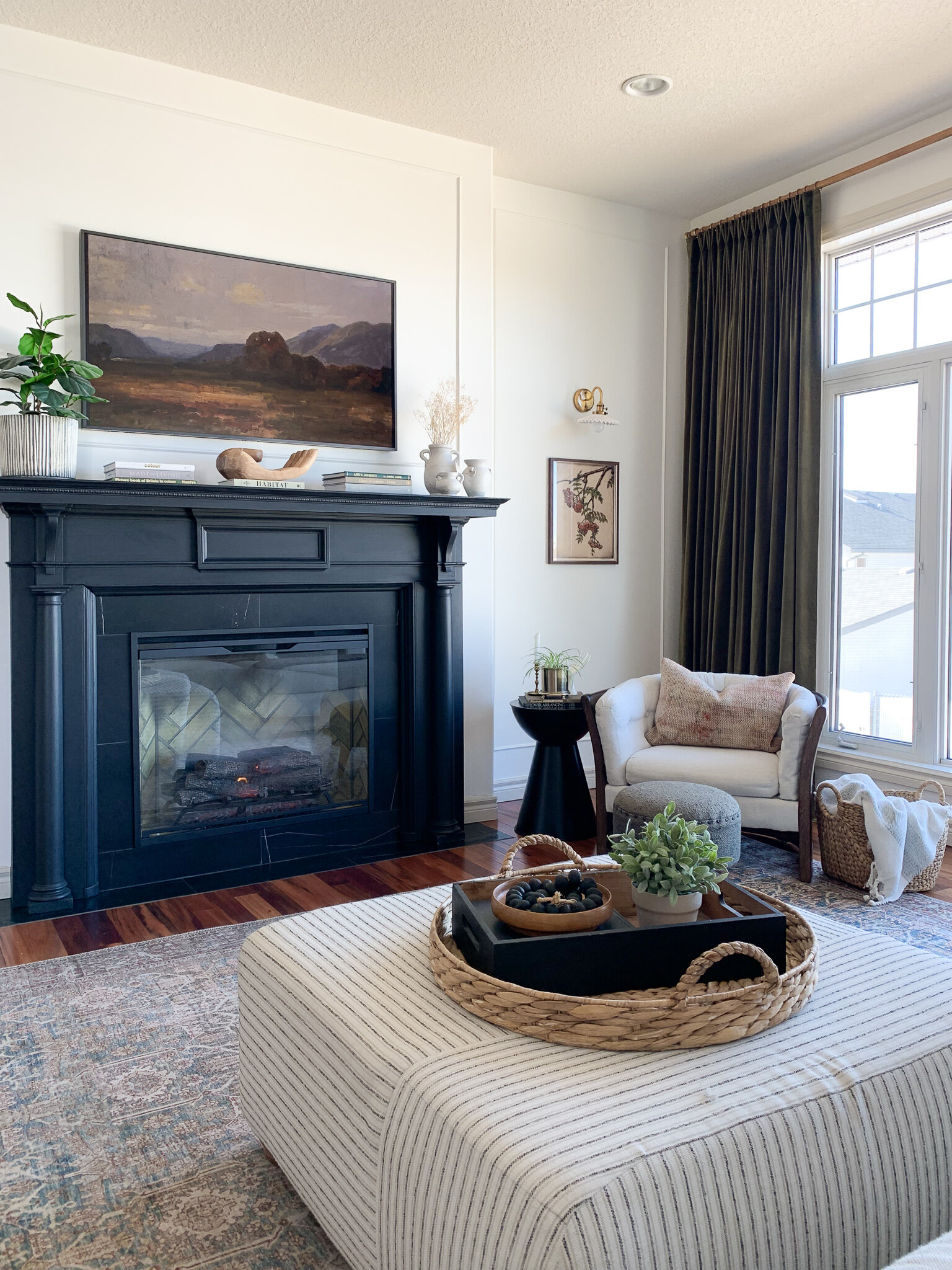 Living room with black antique mantel fireplace with marble surround, green velvet ikea curtains, vintage style loloi rug, striped ottoman, vintage oak chair, brass and ceramic sconce and vintage art