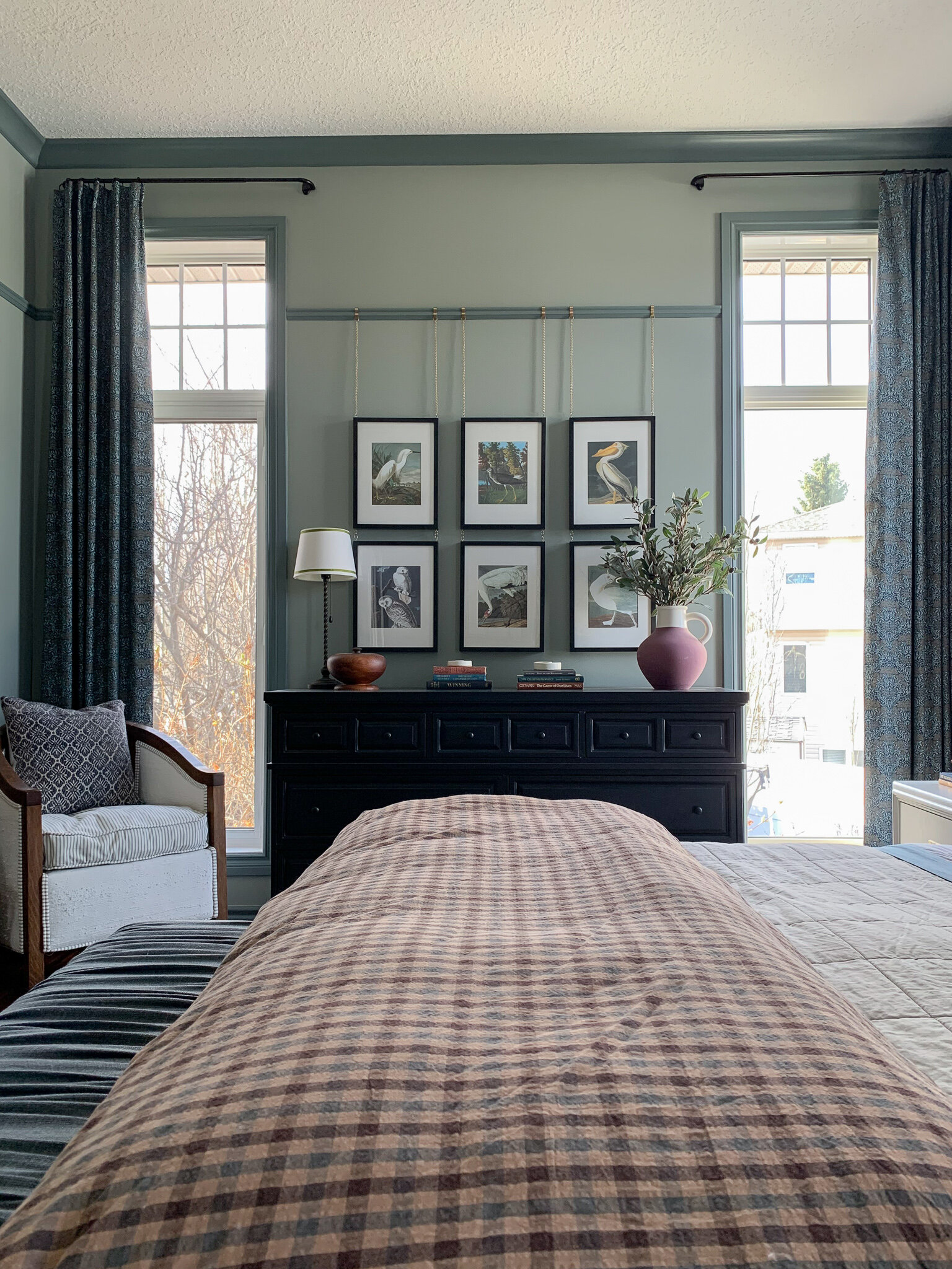 wall with black dresser flanked by two windows with floral curtains, bird art gallery wall hanging from a picture rail, green walls and darker green trim, antique chair in corner, bed in foreground with beige and plaid bedding