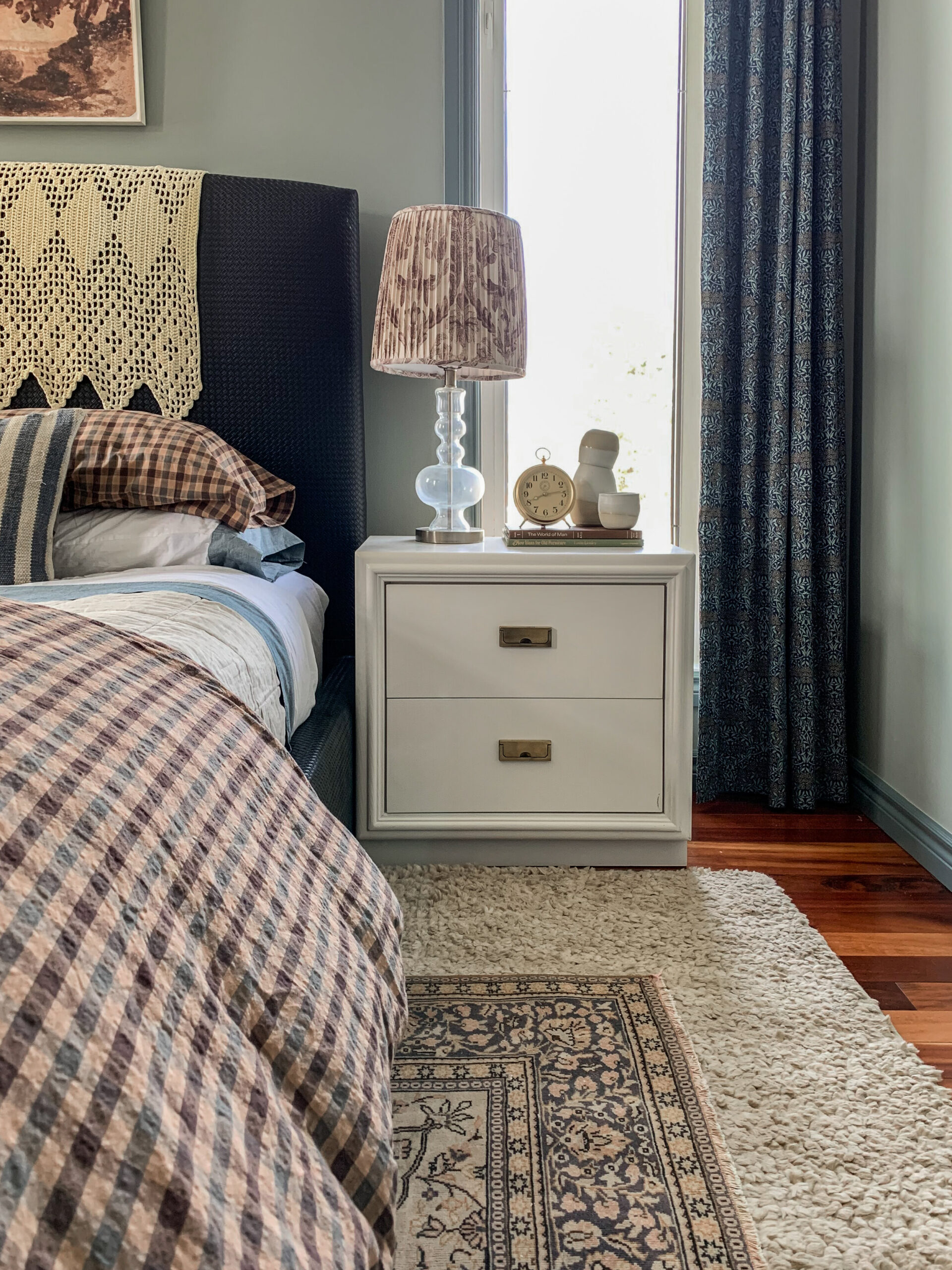 bedside with white nightstands, pleated floral lampshades, floral curtains, green walls with darker green trim, vintage rug and plaid duvet on bed