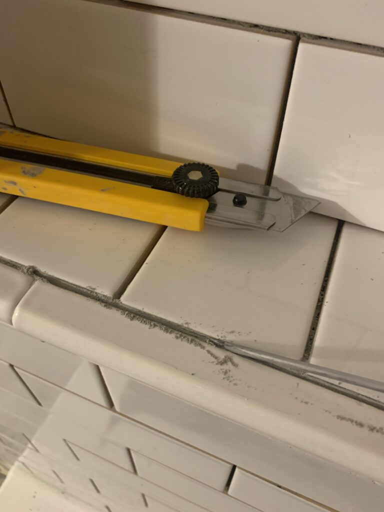 chipping out mortar from between installed subway tiles using a screwdriver and a utility knife