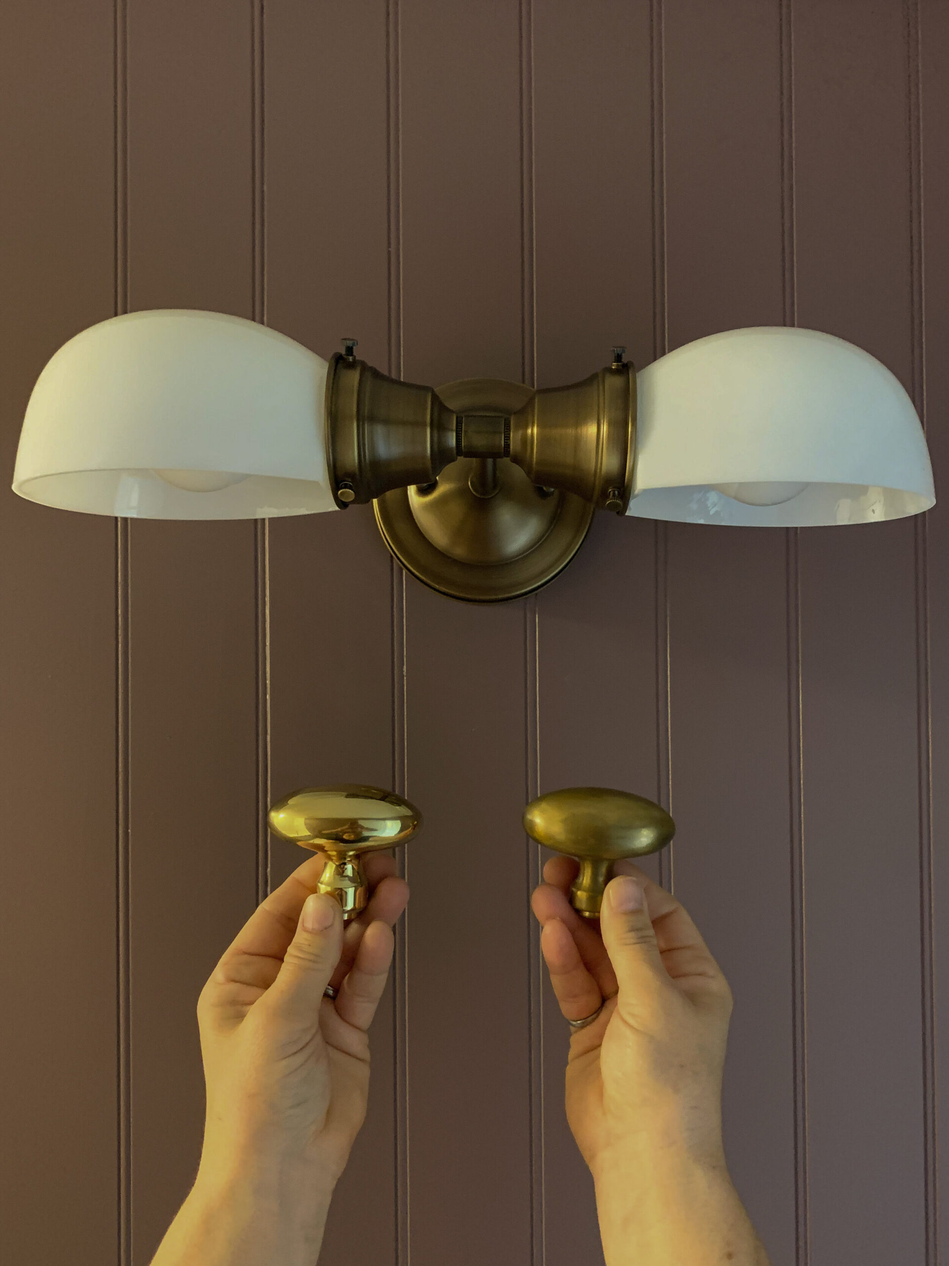 holding up the original laquered brass knob and the unlaquered and aged with vinegar knob to a aged brass light to compare