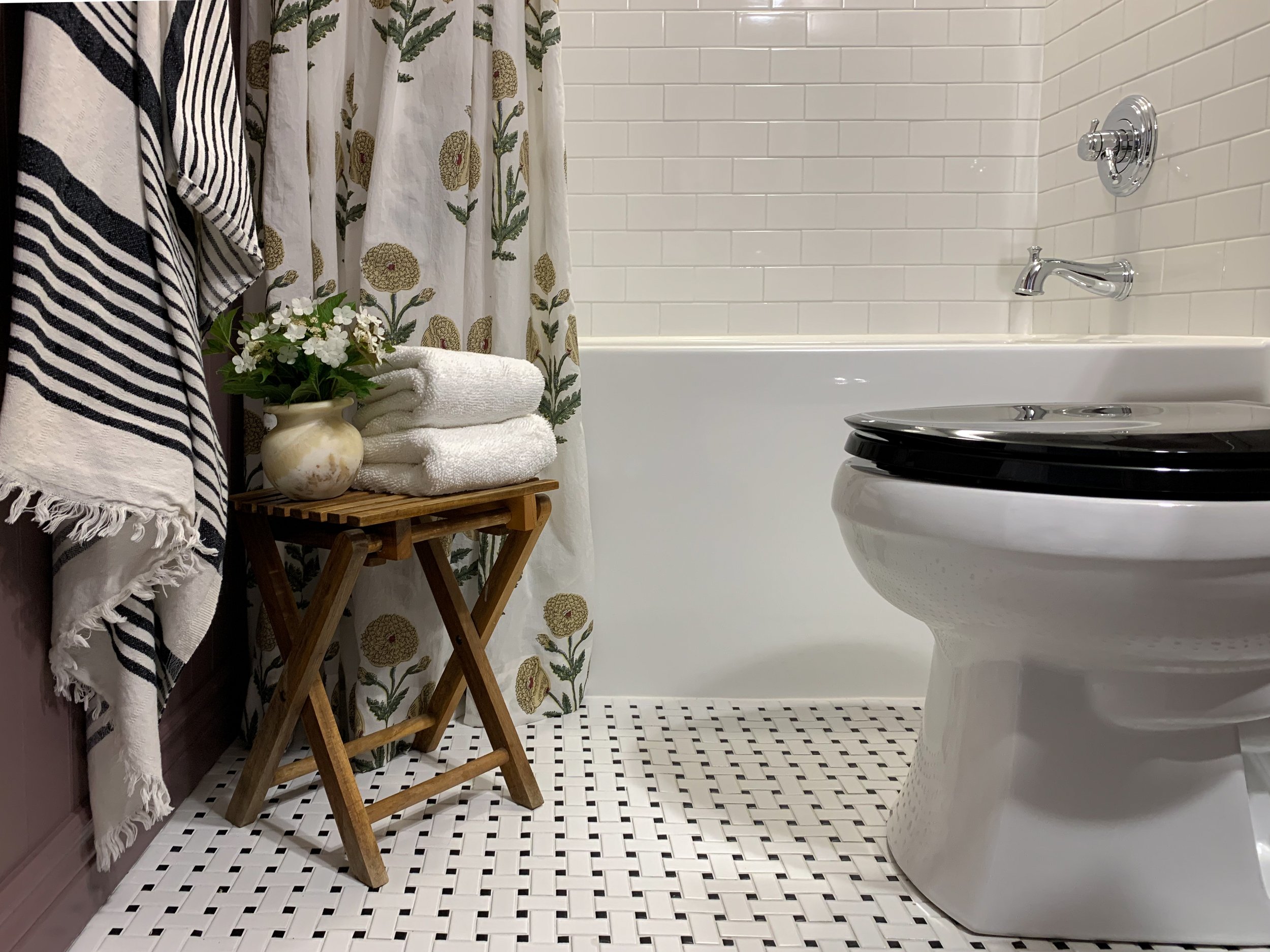 bathroom showing black toilet seat, black and white basketweave floor, white subway tile, purple beadboard, floral shower curtain, vintage stool with white towels and flowers