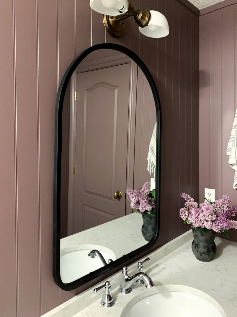 Arch shaped black mirror on a purple beadboard wall with an antique brass light fixture above, a white quartz counter and chrome vintage style faucet