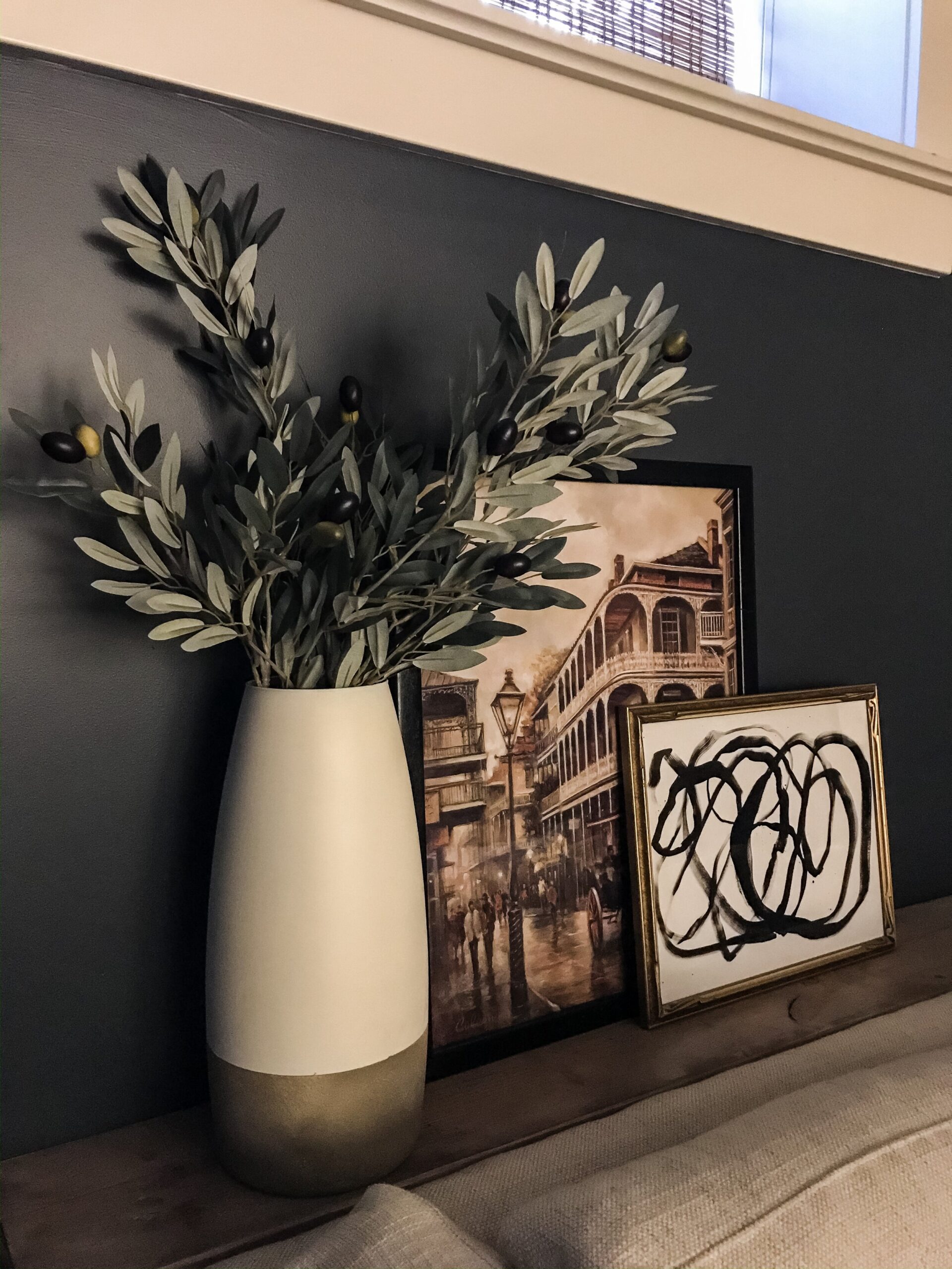 blue wall with two peices of art layered on a shelf with a vintage painting and abstract sketch, and a vase with greenery