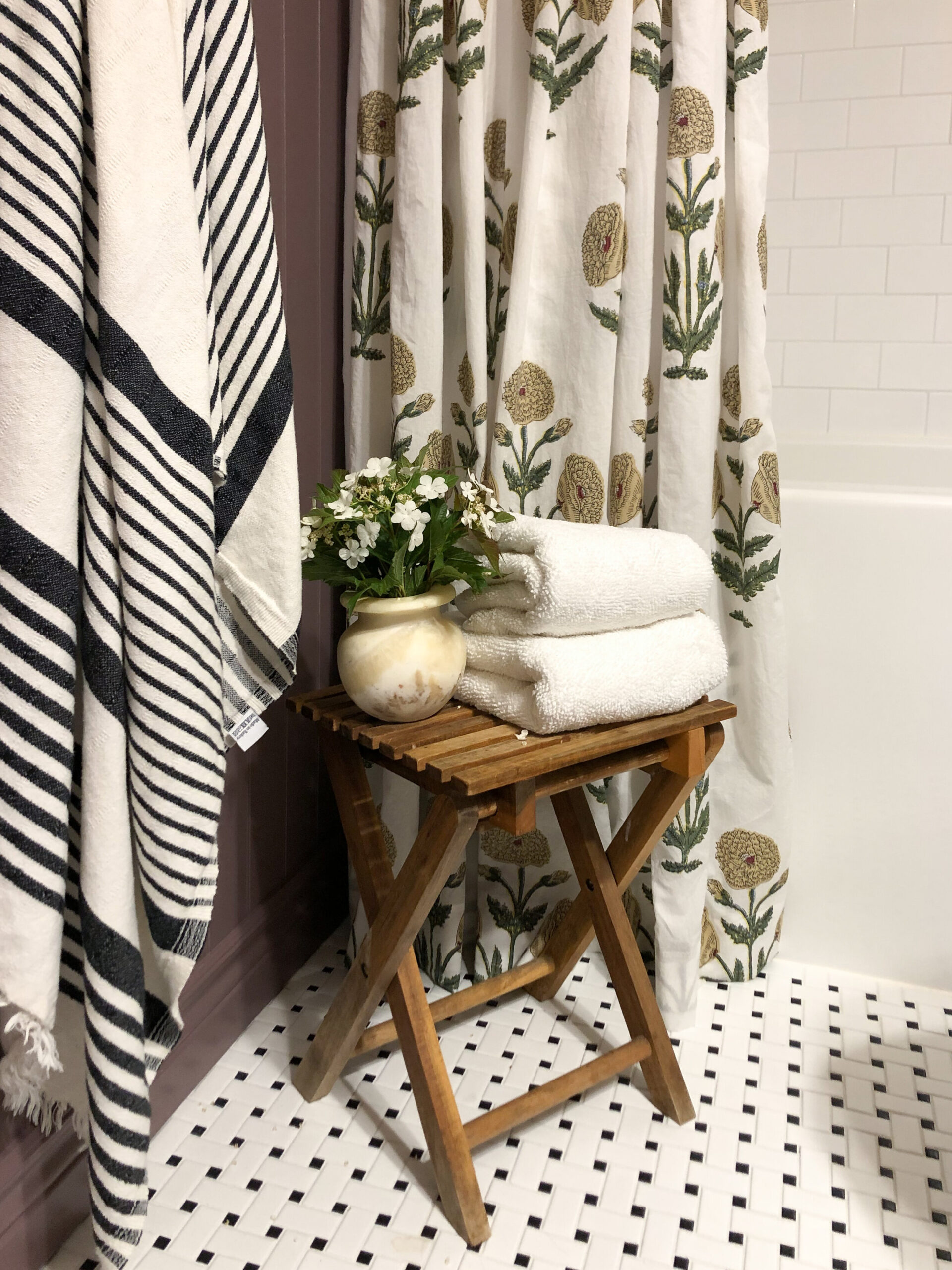 Block print floral DIY shower curtain with a black and white striped towel hanging on a hook, purple beadboard walls, white subway tile, and a vintage wood stool with towels and flowers 