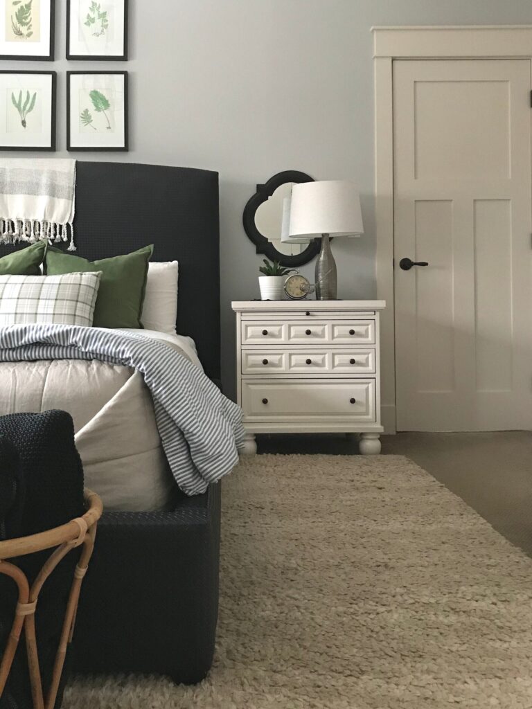 picture of a white nighstand alongise a lack bed with beige and blue striped bedding and green pillows, and a lamp and styling items on top