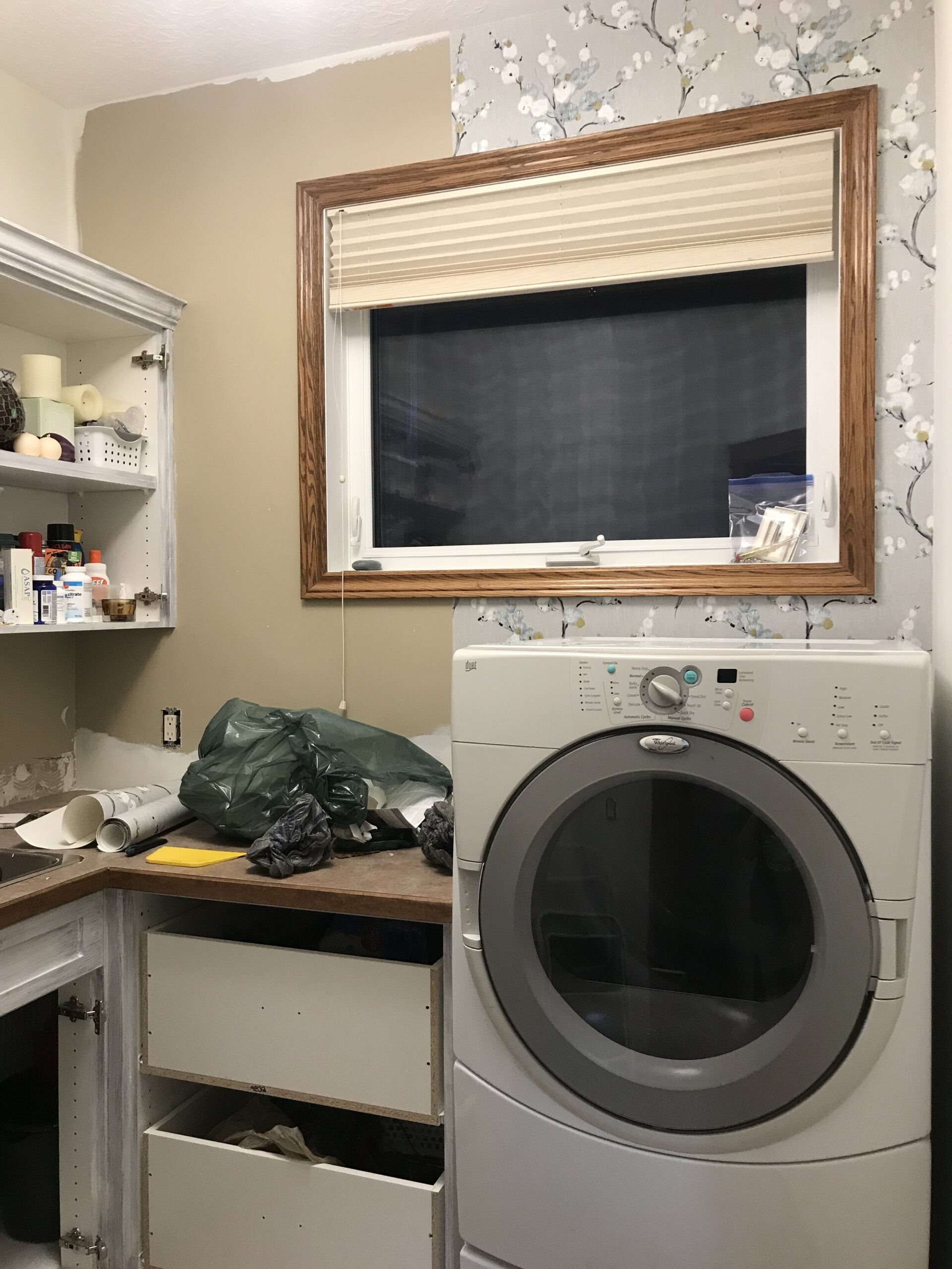 laundry room under construction with half wallpapered wall and doors all removed from cabinets, a dryer and a window