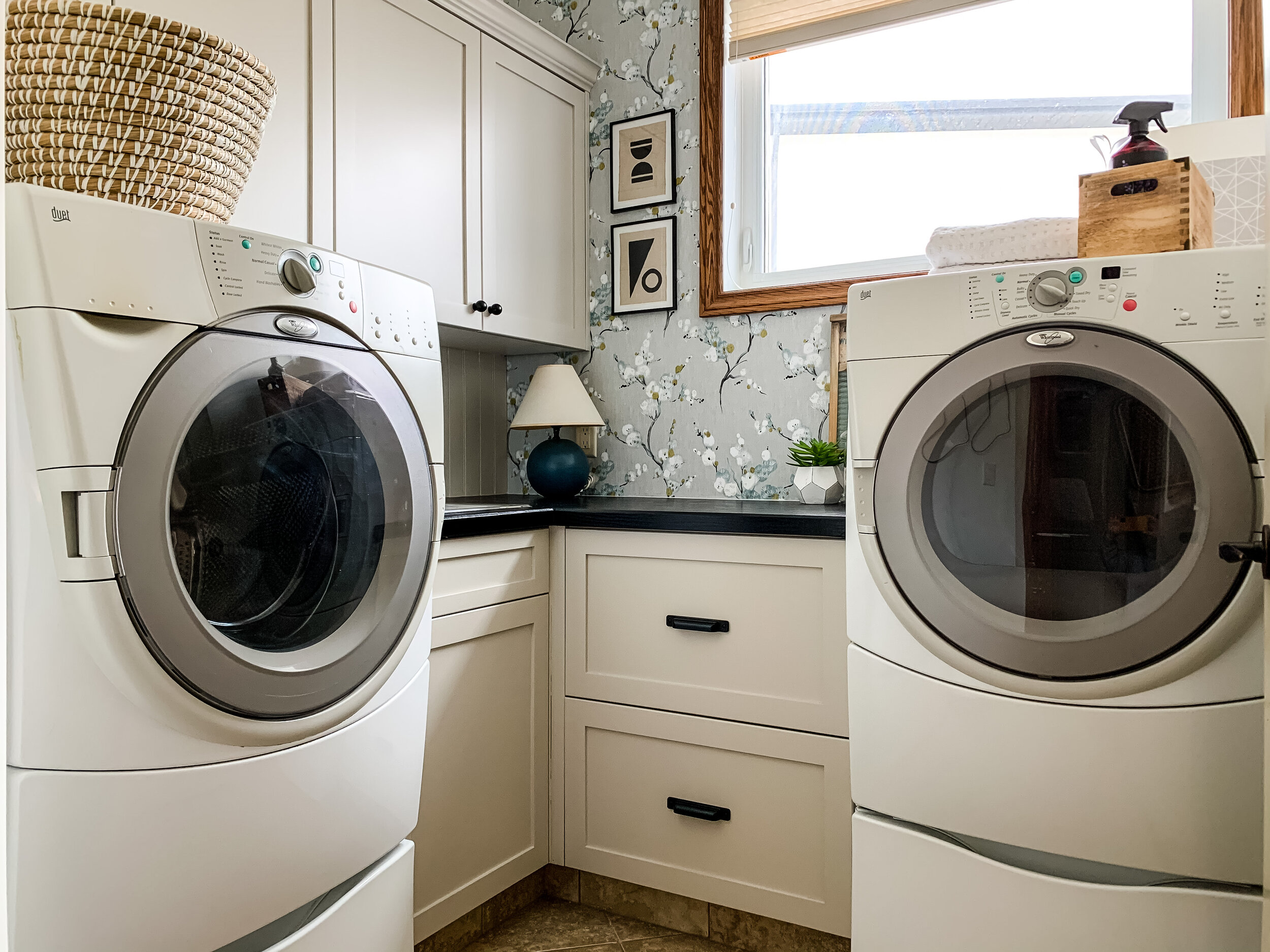 laundry room with beige cabinets with black counters, floral grey wallpaper, small blue lam, modern art, some styling pieces on counter