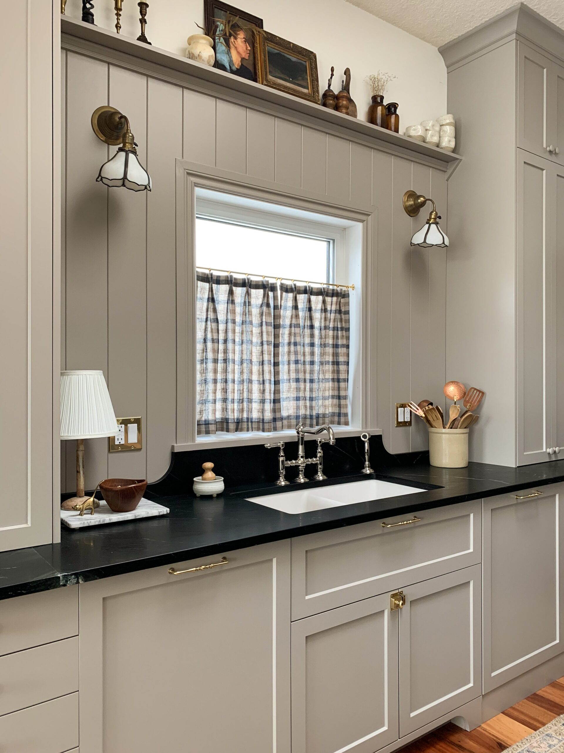 kitchen sink area with countertop cabinets flanking the sink, soapstone counters with curved stone backsplash up to a window, paneled wall with shelf and vintage pendants, cafe curtain on window