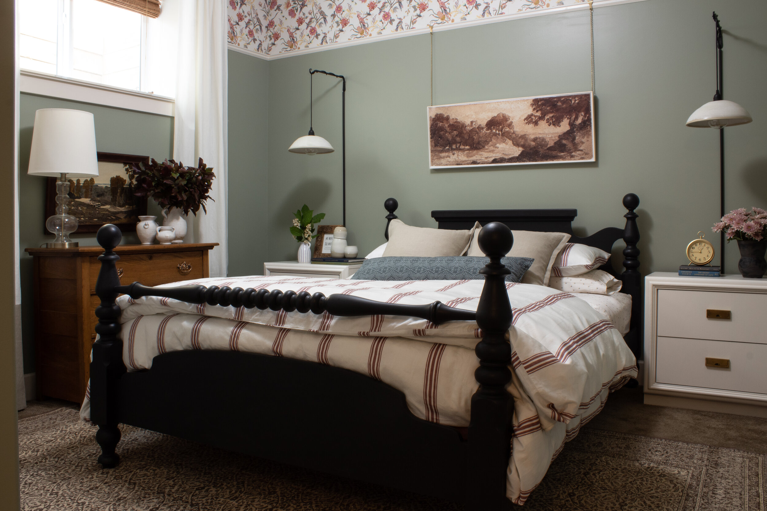 bedroom with green walls and picture rail trim with floral wallpaper above the picture rail, red striped bedding, black antique bed, white nightstands and a brown vintage rug
