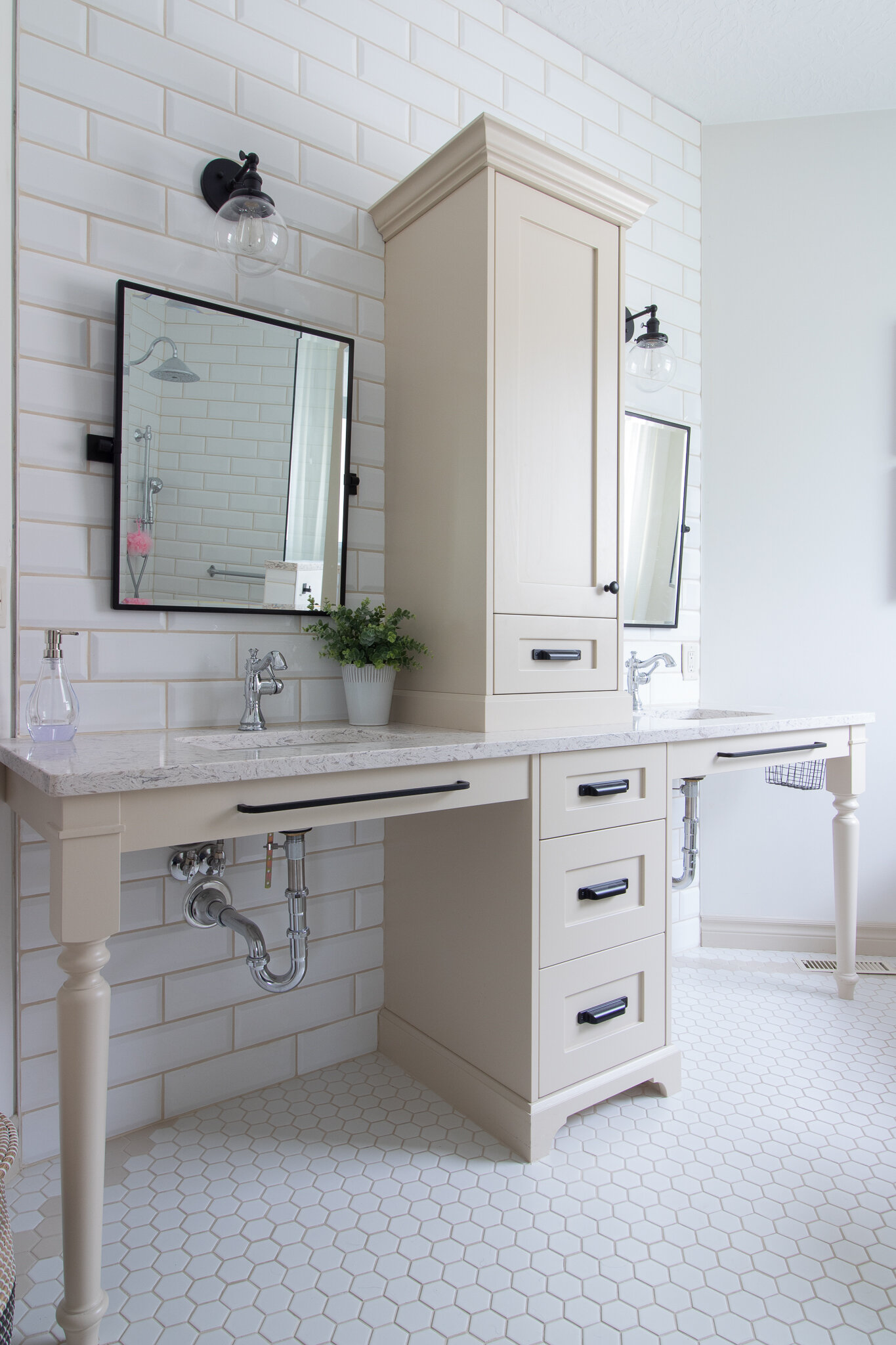 Wheelchair friendly bathroom vanity with two sinks, a central cabinet tower, tiled subway wall, beige cabinetry and quartz counters, chrome single handle fixtures, beveling mirrors and black globe light fixtures with hex tile on floor with border