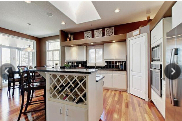 white kitchen with black granite counters and black backsplash, ledge over top of cabinet uppers, corner pantry, window, raised eating bar and wine rack on angled island