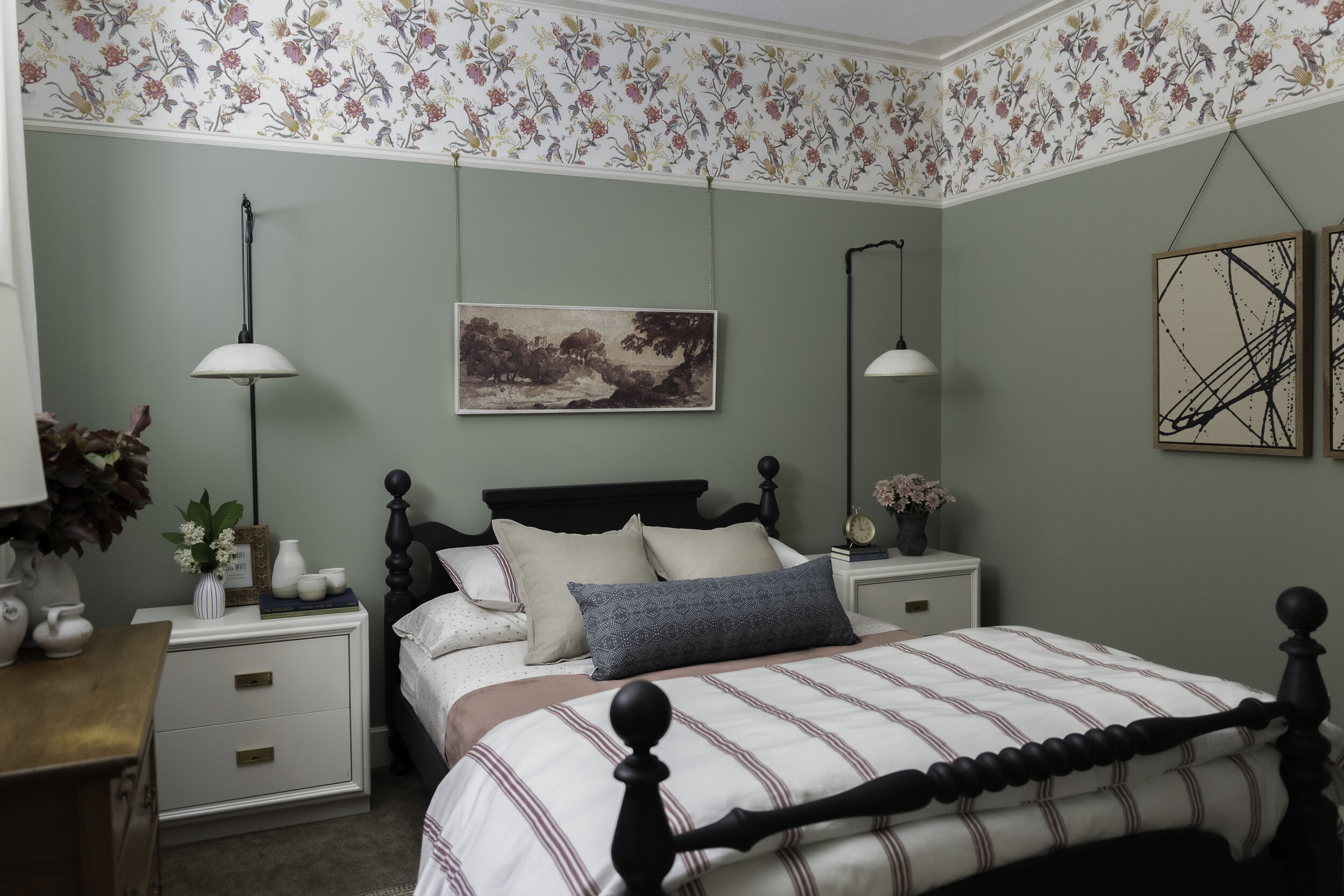 green bedroom with wallpaper at top of wall, red striped bedding, white modern nightstands, hanging ceramic wall sconces, vintage rug and black antique bed and antique wood dresser