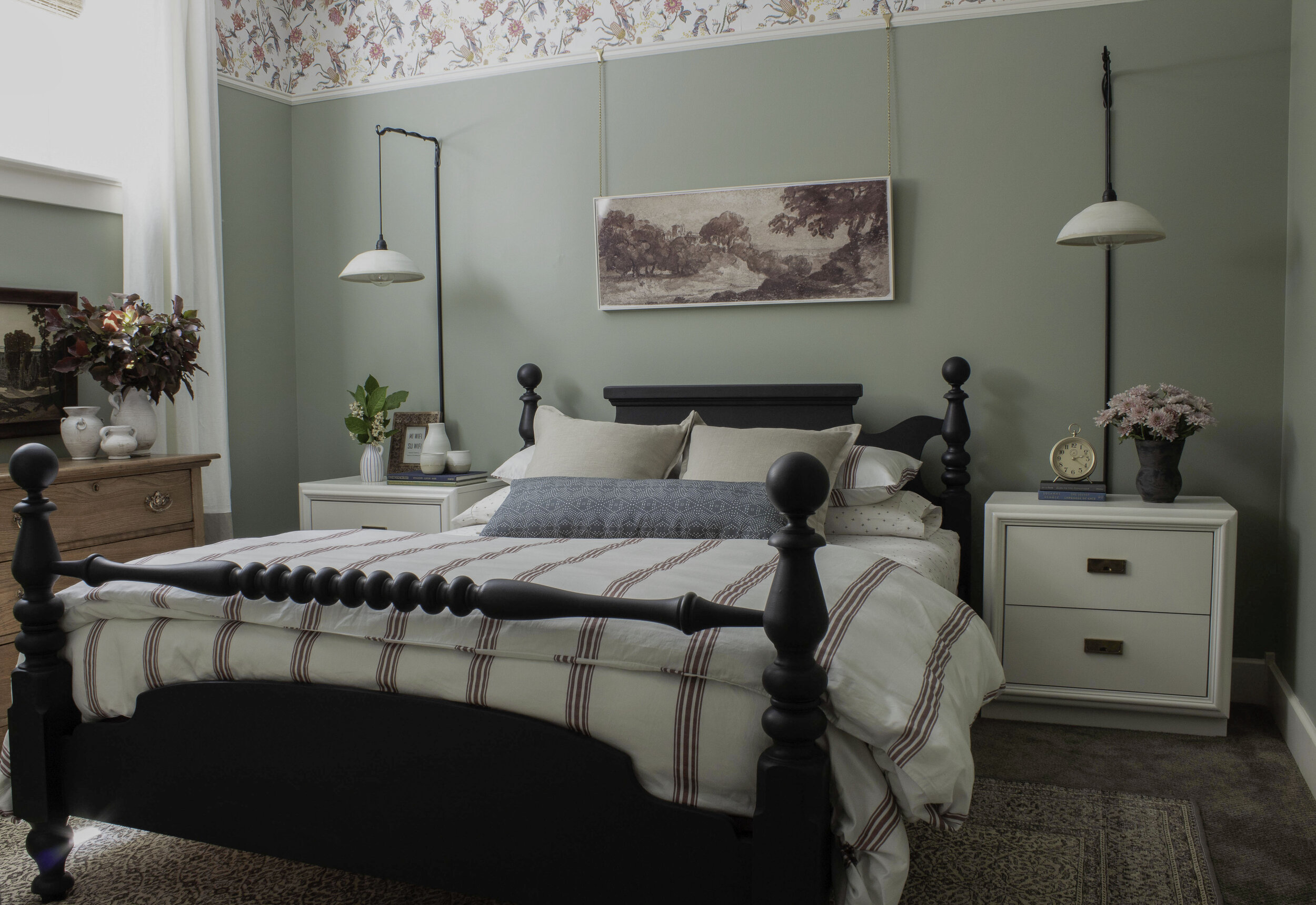 green bedroom with wallpaper at top of wall, red striped bedding, white modern nightstands, hanging ceramic wall sconces, vintage rug and black antique bed and antique wood dresser