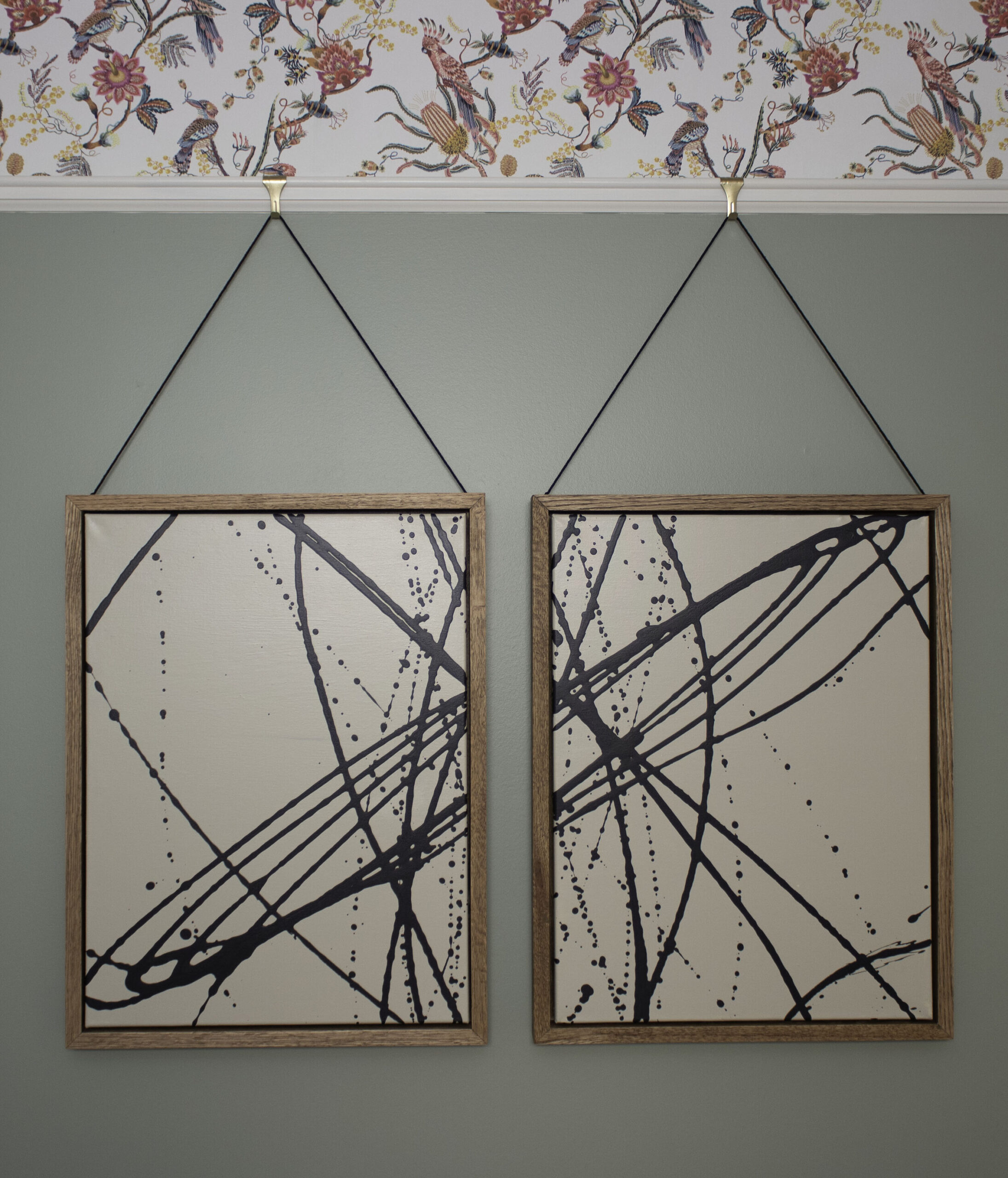 black and beige abstract art with oak wood frames hanging by strings from a chair rail on a green wall