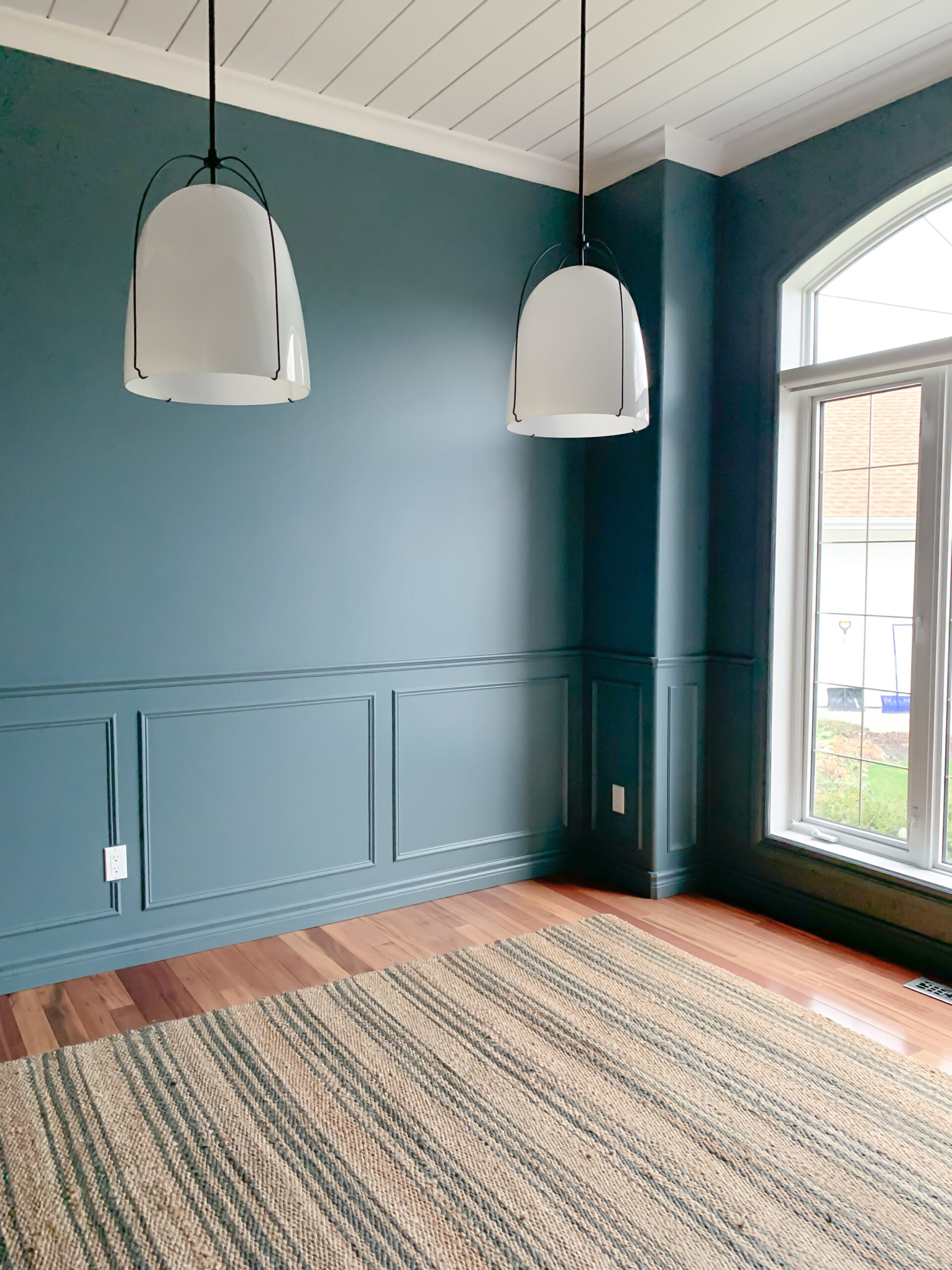 empty room with box moulding and chair rail on the bottom part of the room, blue walls, wood floors and jute striped rug, white glass pendants and white shiplap ceiling
