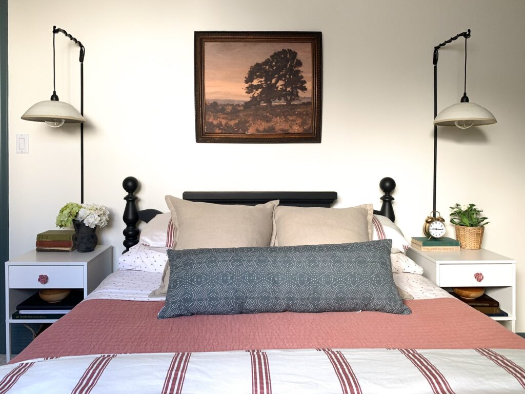 veiw of bed with vintage art above, vintage black painted bed, pink striped bedding, ceramic wall hanging sconces and white ike nightstands