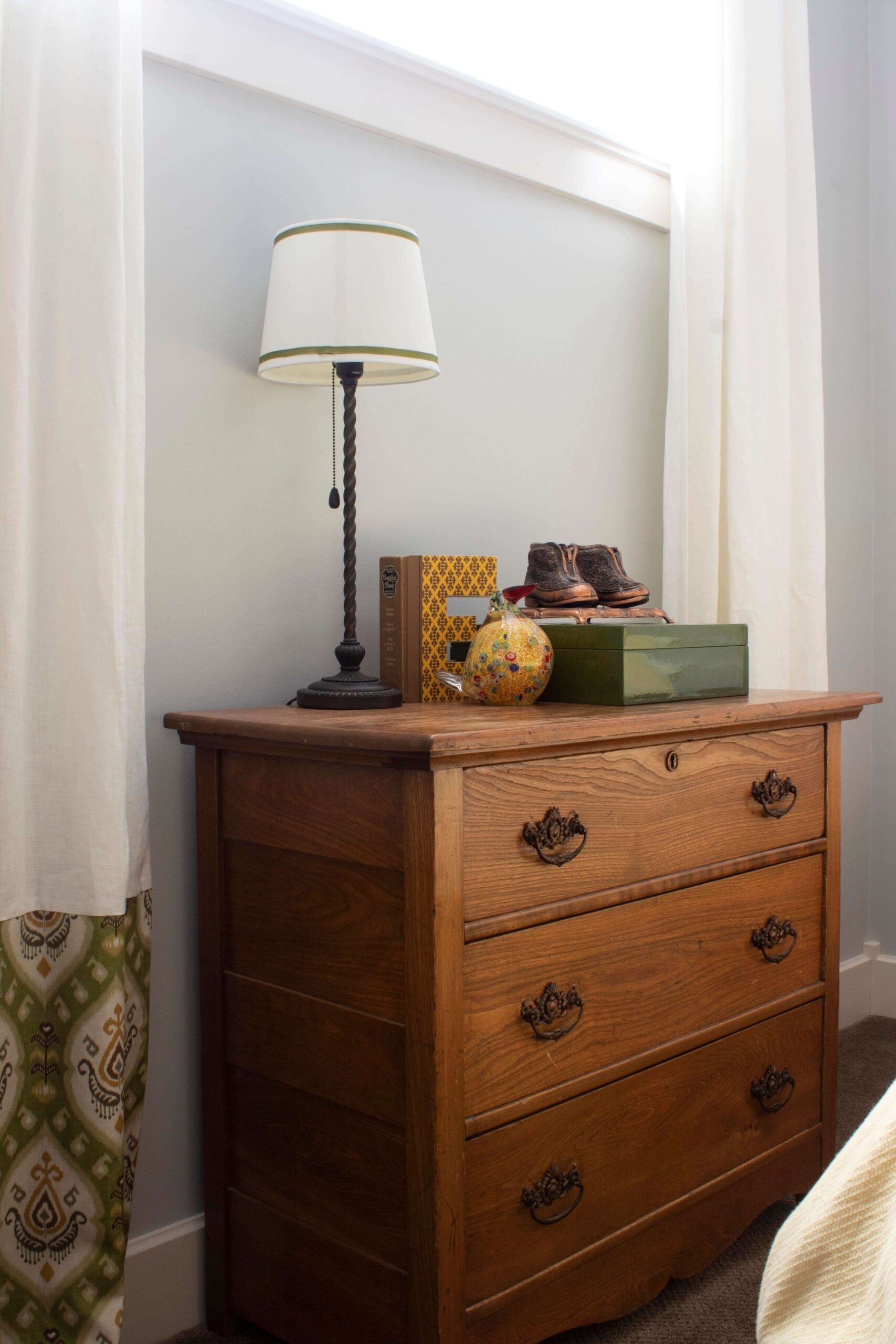 Antique dresser styled with various items and a lamp, grey walls and window above, flanked by white curtains