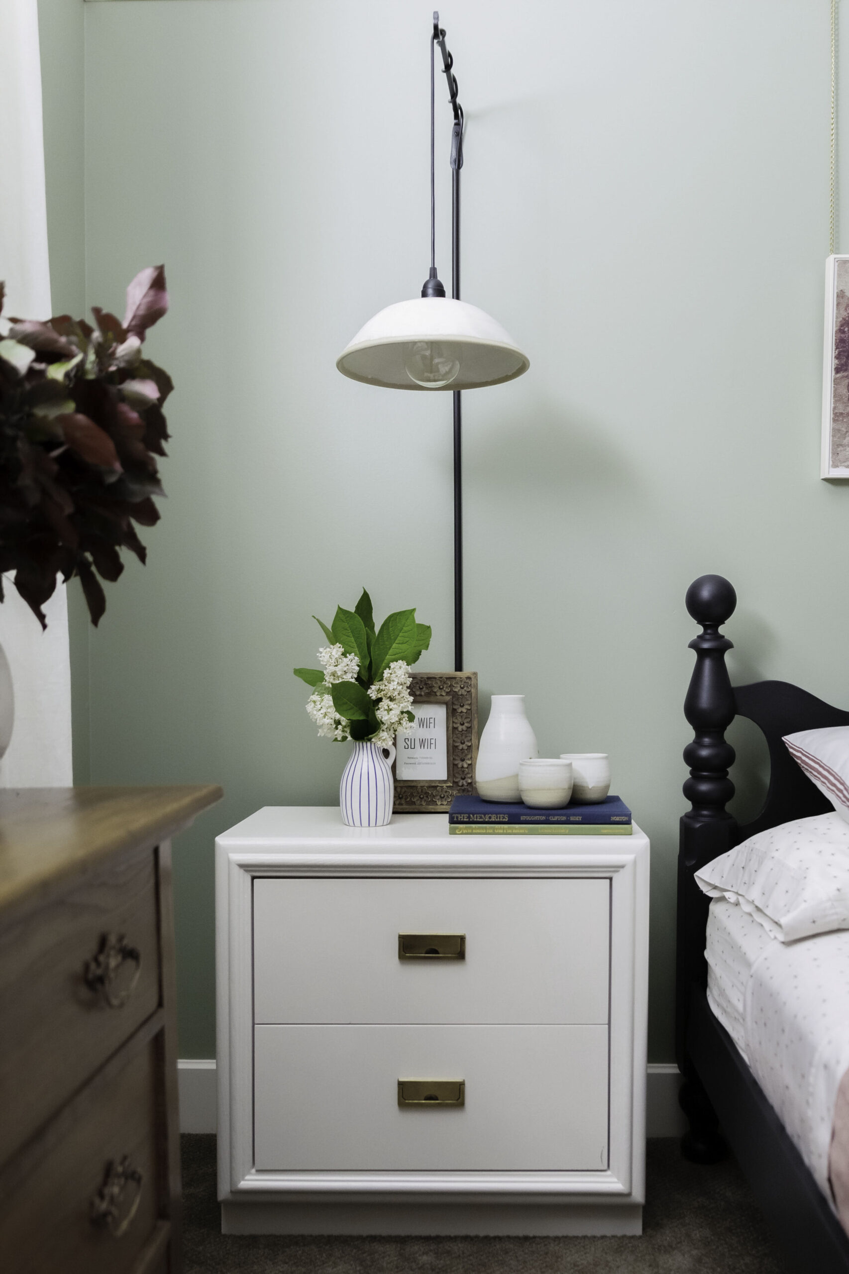 white modern nightstand with black spindle bed beside and with ceramic wall sconces hanging above, with flowers and books/cermaics styled on nightstand