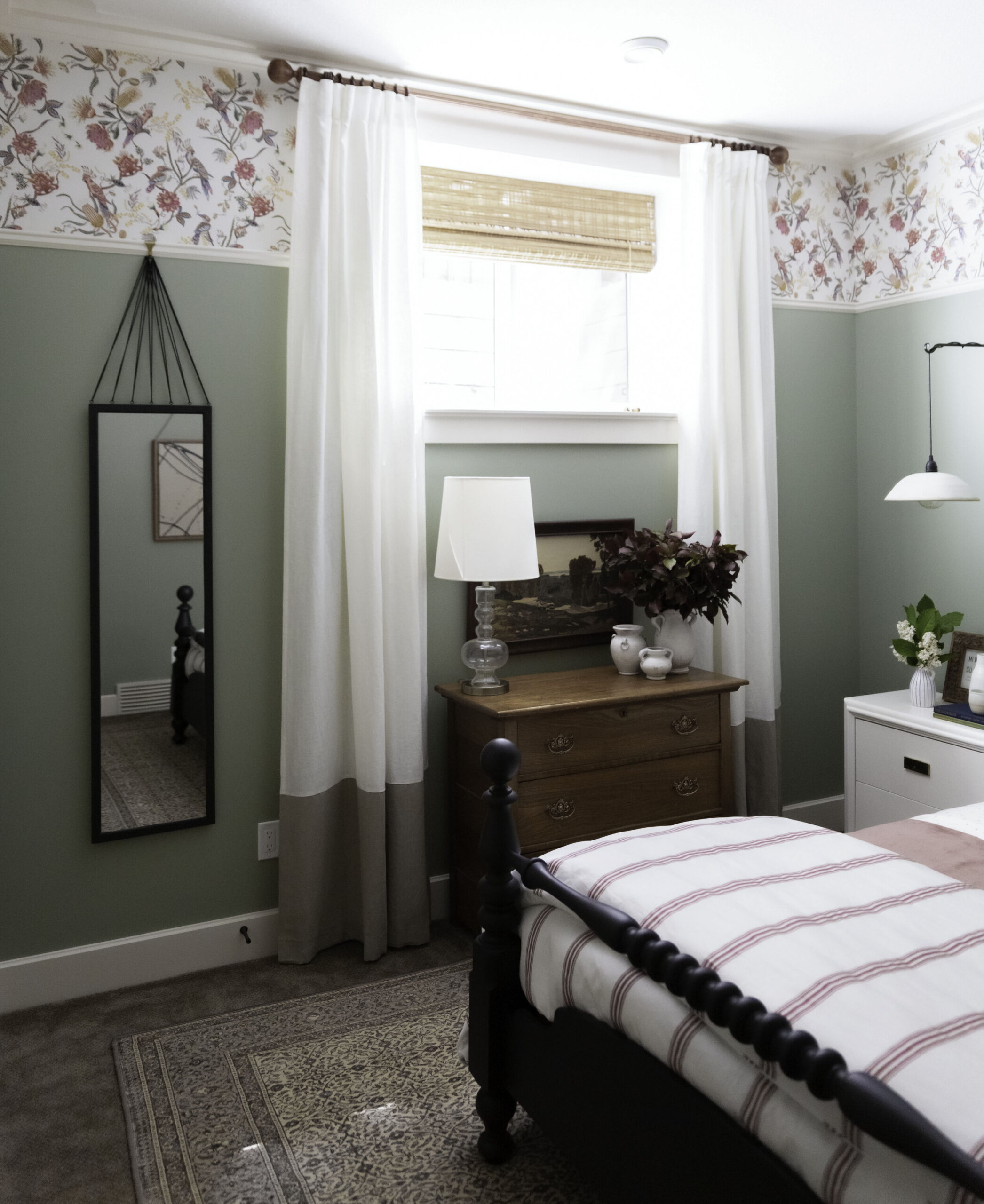 green bedroom with white curtains with beige colour blocking, mirror with strings hanging from picture rail with floral wallpaper above the picture rail, window with antique wood dresser below, black bed with pink striped bedding