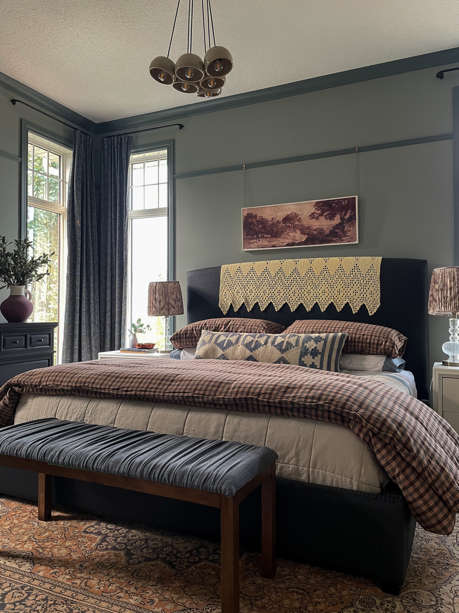 Bedroom with green walls with darker green trim, picture rail, two windows and floral curtains, beige and checkered bedding, afghan over black headboard on bed, art over bed and white nightstands with glass lamps with pleated floral shades