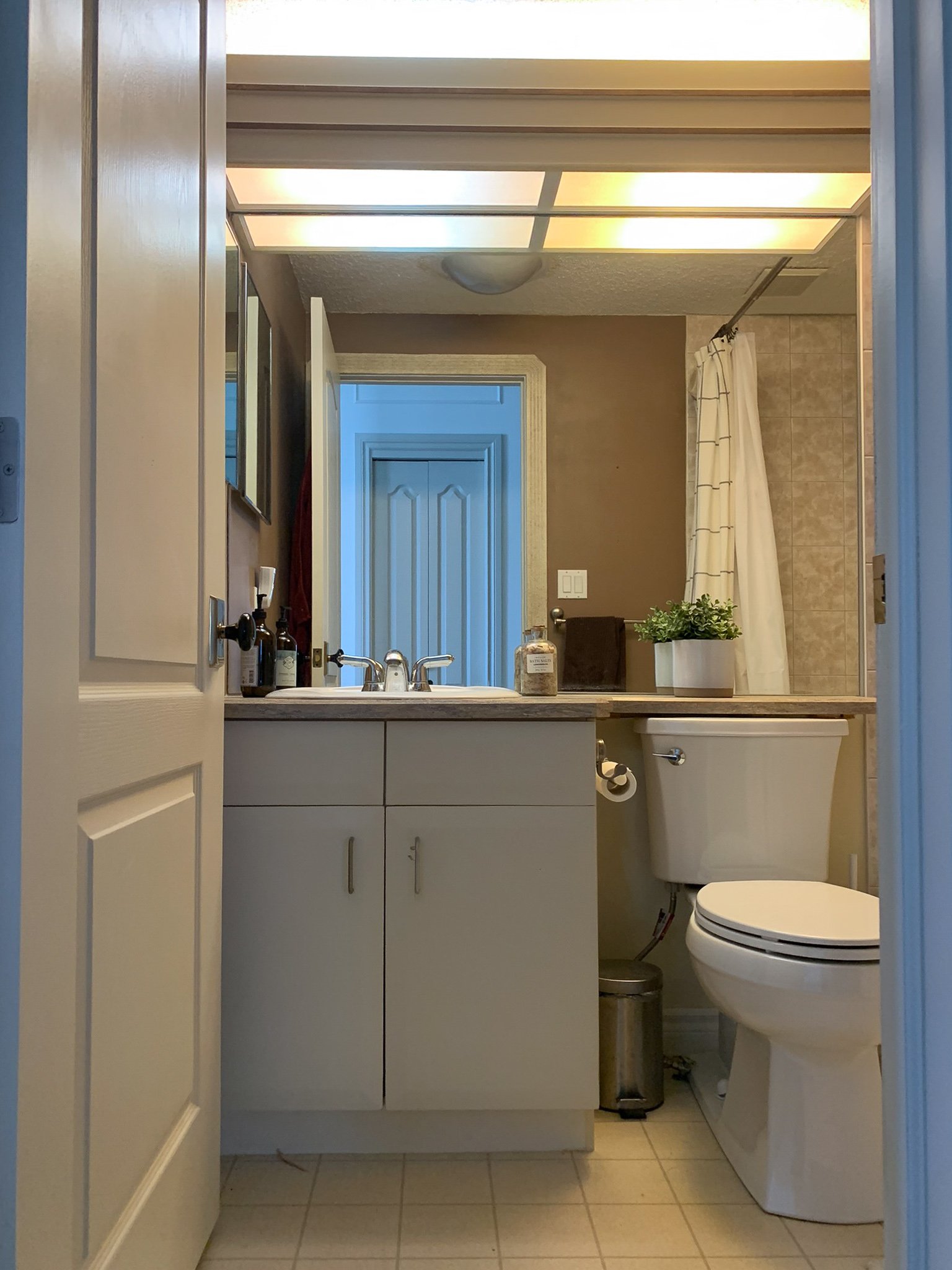Bathroom before with flourescent light, frameless mirror, flat front white cabients, laminate counters and linoleum floor