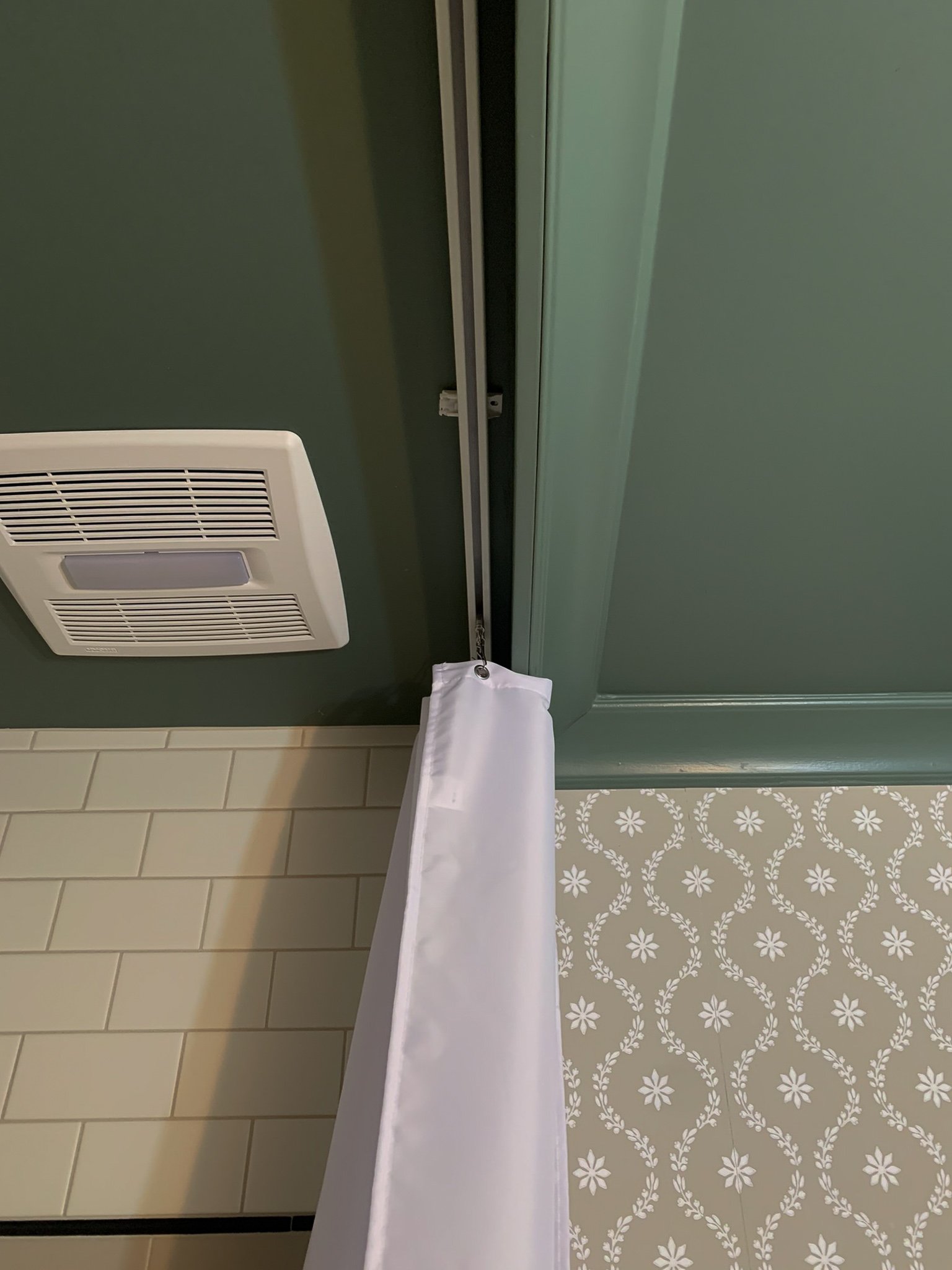 shower curtain installed on a ceiling mounted curtain track with green ceiling and crown moulding, subway tile shower and floral wallpaper in background